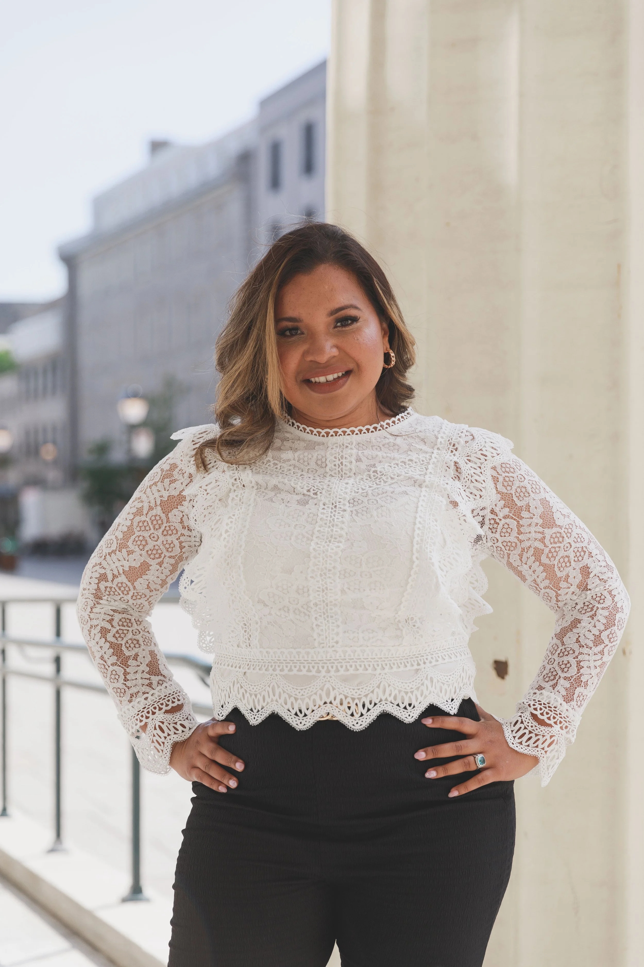 Une femme souriante pose en extérieur, portant une blouse blanche en dentelle avec des manches longues et un col rond, et un pantalon noir.