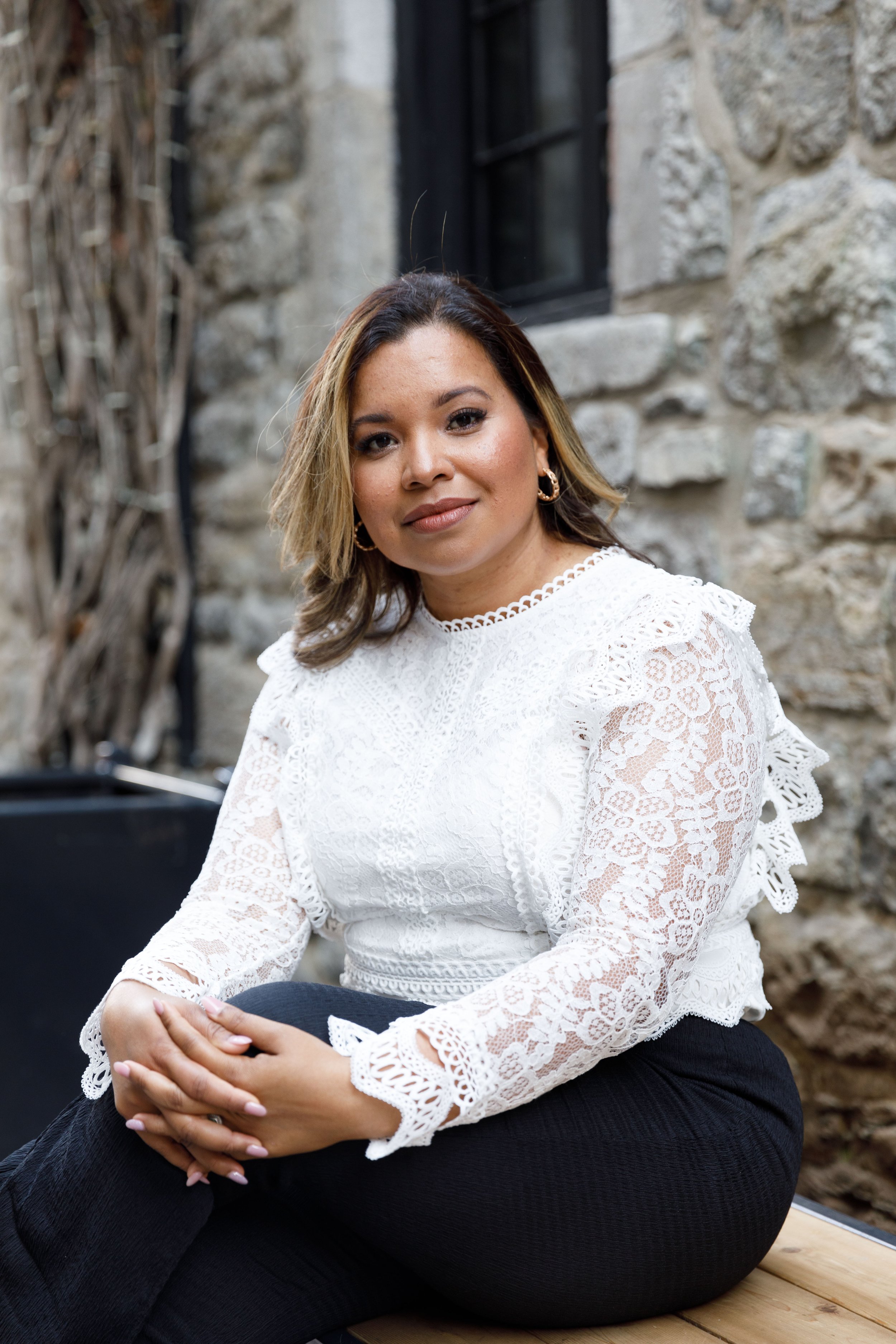 Une femme assise en extérieur contre un mur en pierre, portant un haut blanc en dentelle et des pantalons noirs, souriant légèrement, avec des cheveux mi-longs bruns et bouclés, portant des boucles d'oreilles en or.