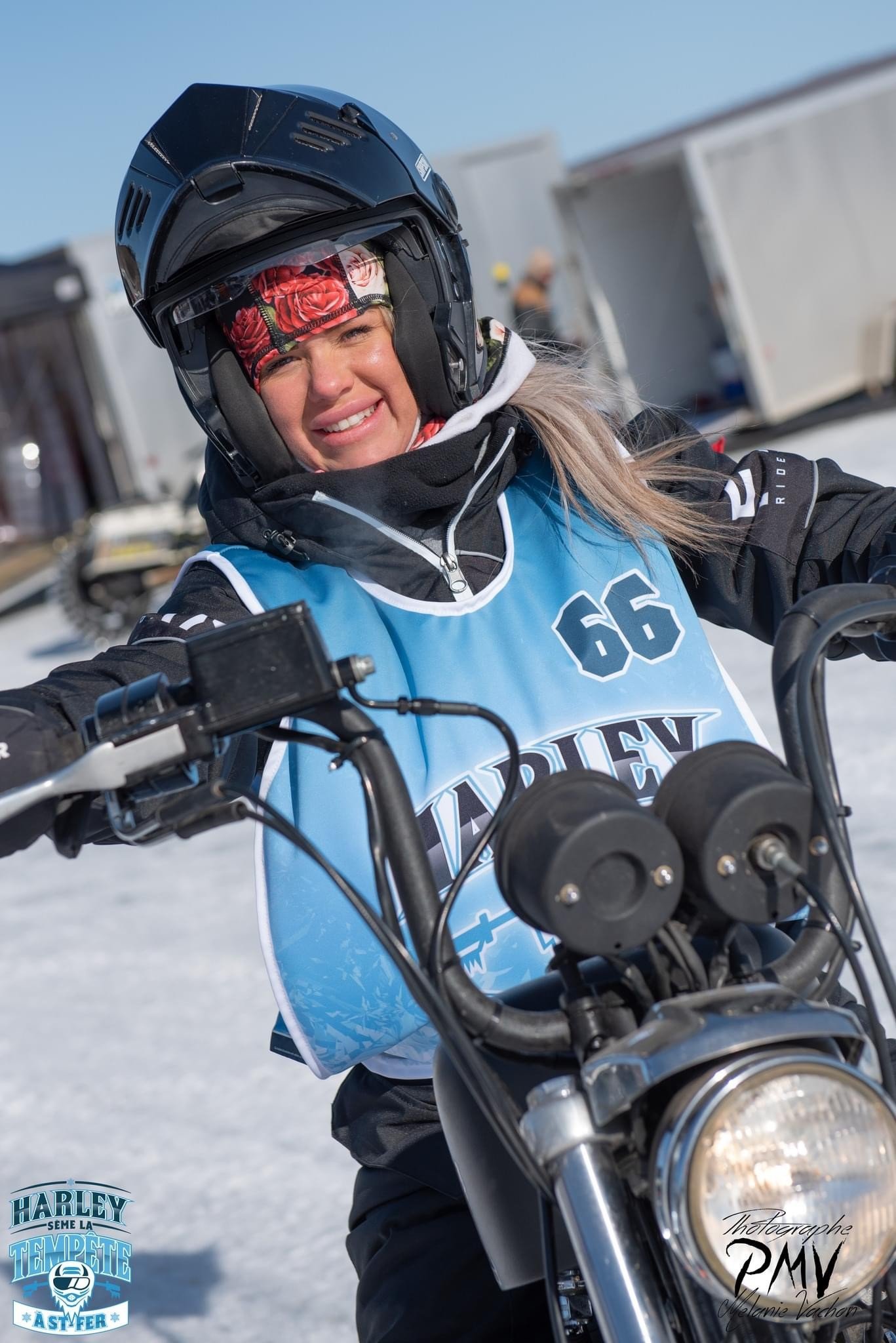 Une jeune femme souriante portant un casque de moto et un vêtement de sport, assise sur une moto en plein air enneigé, avec des structures en arrière-plan.