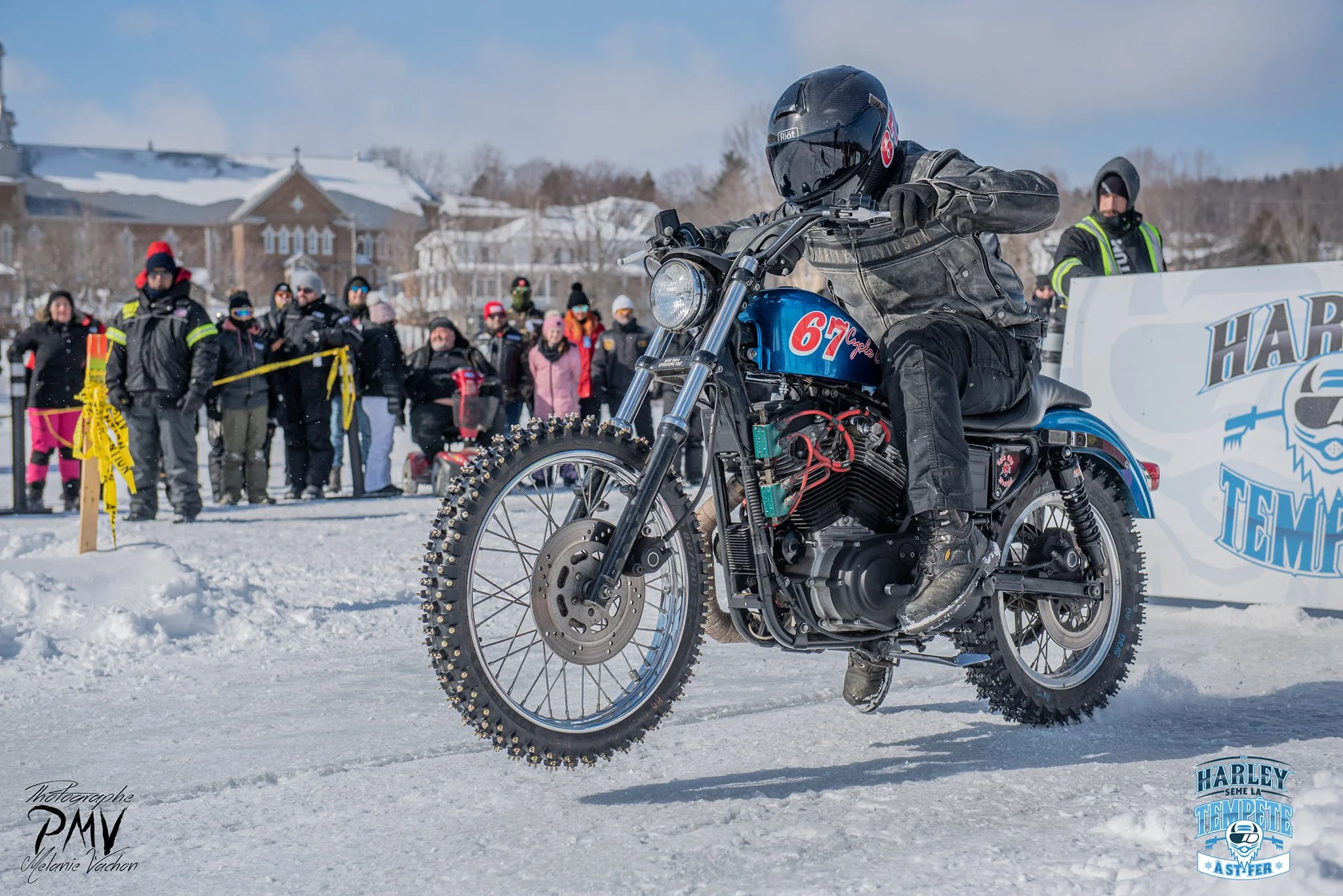 Un motard en uniforme noir avec casque noir conduit une moto sur la neige avec un groupe de spectateurs en arrière-plan. Il y a un panneau d'événement "Harley Tempête" à droite.