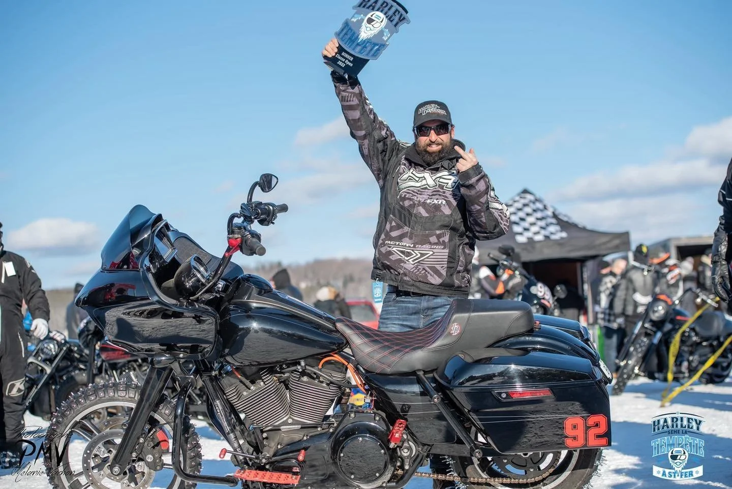 Un homme souriant, portant une veste de moto, célèbre sa victoire en levant un trophée, à côté d'une moto de couleur noire avec le nombre 92 sur la côté, en plein air avec d'autres personnes et motos en arrière-plan.