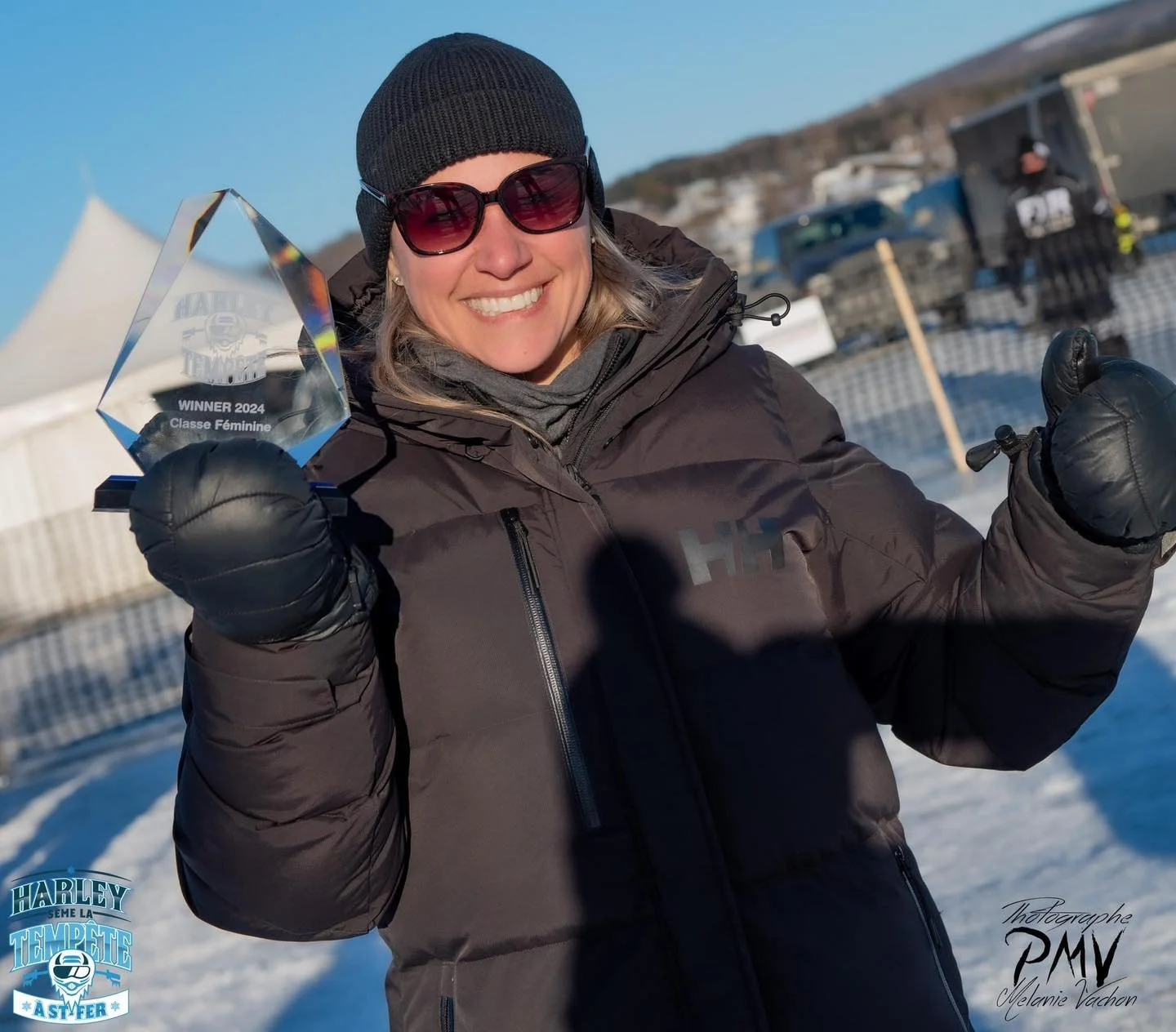 Femme souriante portant un bonnet noir et des lunettes de soleil, tenant un trophée en verre indiquant qu'elle a gagné en 2024 dans la catégorie féminine, en plein air avec neige et voitures en arrière-plan.