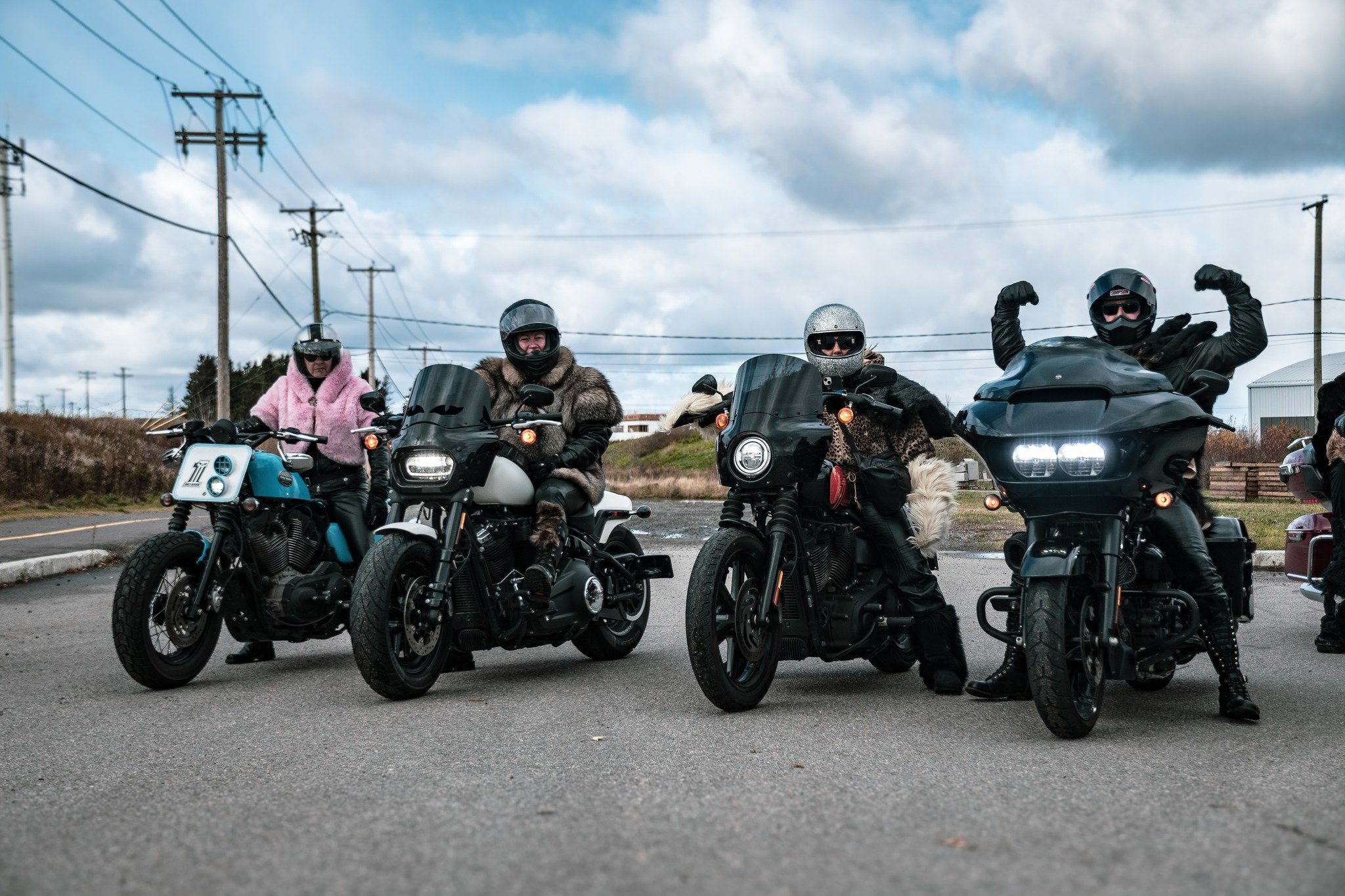 Quatre motards en vêtements chauds et casques, posant avec leurs motos sur une route, sous un ciel nuageux, avec des lignes électriques en arrière-plan.