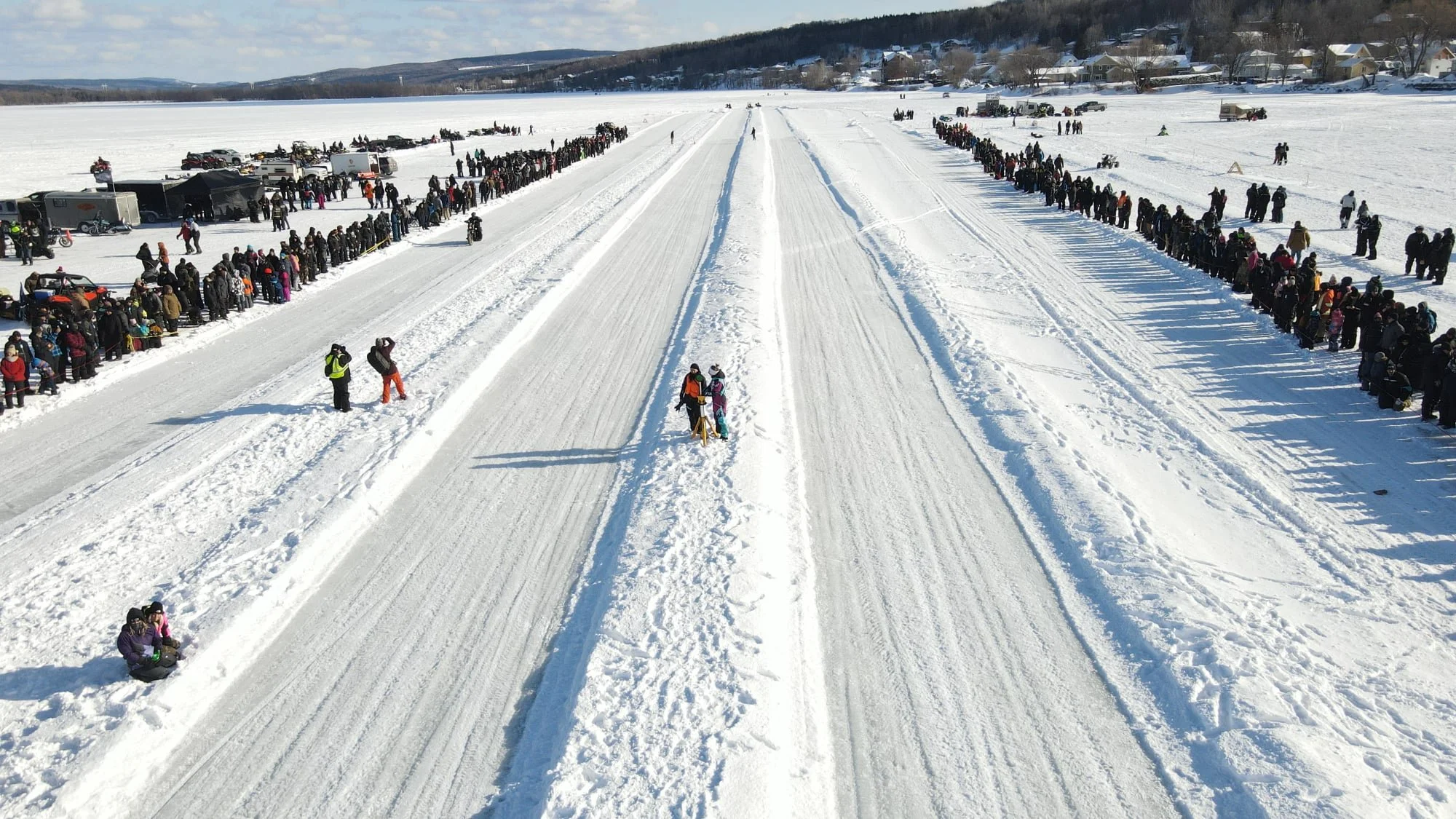 Une masse de personnes attendant dans une file le long d'une rivière gelée et enneigée, avec des voitures stationnées à l'arrière-plan, dans un paysage hivernal.