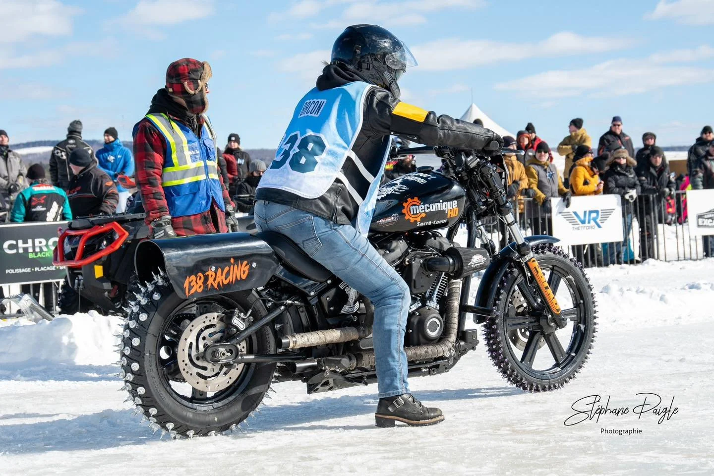 Un motocycliste en tenue de course, portant un casque noir, est prêt à démarrer sur une piste enneigée, avec un public en arrière-plan derrière une barrière.