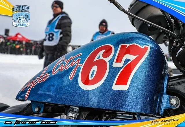 Une moto avec le numéro 67 et l'inscription 'Cycle City' sur le réservoir, des personnes en vêtements d'hiver et un drapeau au fond dans un environnement enneigé