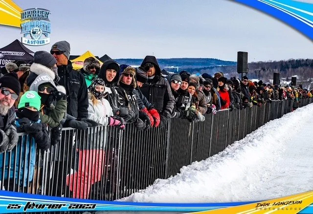 Groupe de spectateurs vêtus de vêtements d'hiver, derrière une barrière en métal, regardant un événement en plein air sur un terrain enneigé.