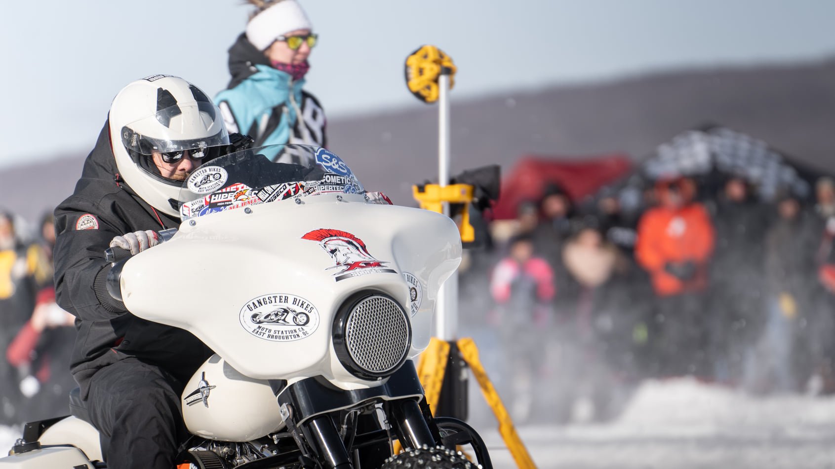 Un motard en combinaison noire et casque blanc, portant des lunettes de soleil jaunes, sur une moto de style vintage avec des stickers, devant un public lors d'une course ou démonstration sur neige.