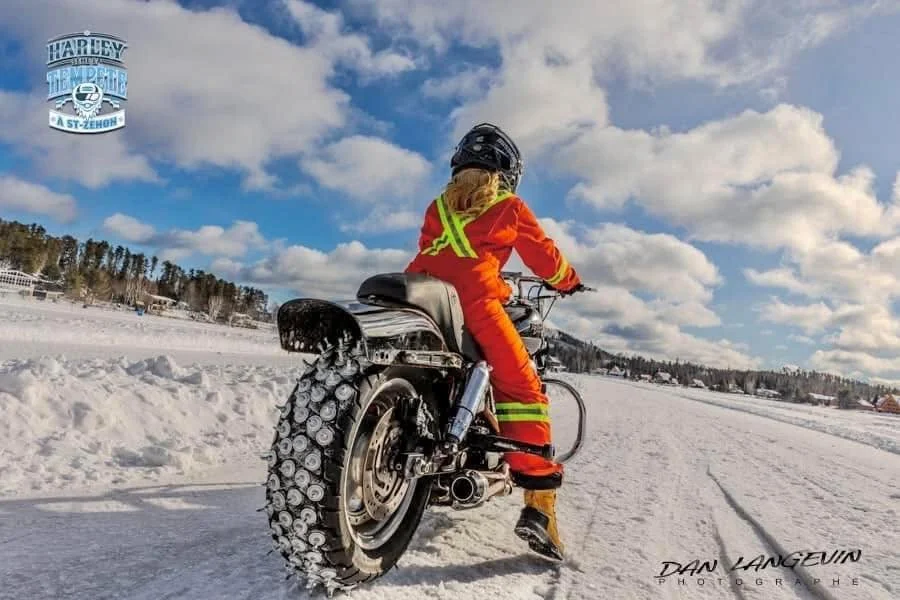 Une fille en tenue orange et casque noir roule en moto sur la neige, avec des pneus à crampons, dans un paysage enneigé sous un ciel partiellement nuageux.
