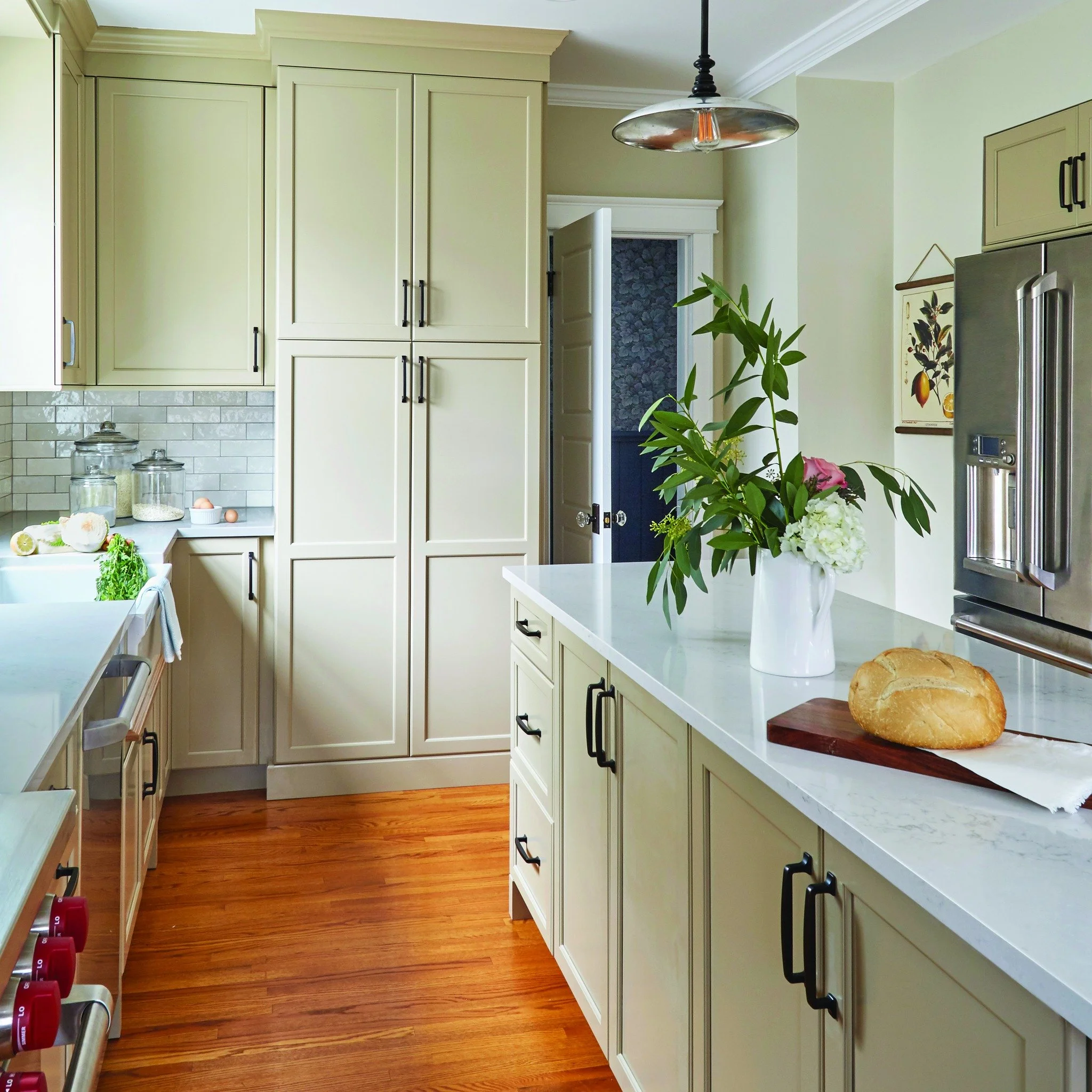 A bright kitchen with white cabinets, a marble countertop with a bouquet of flowers and a loaf of bread, hardwood floor, and a stainless steel refrigerator.
