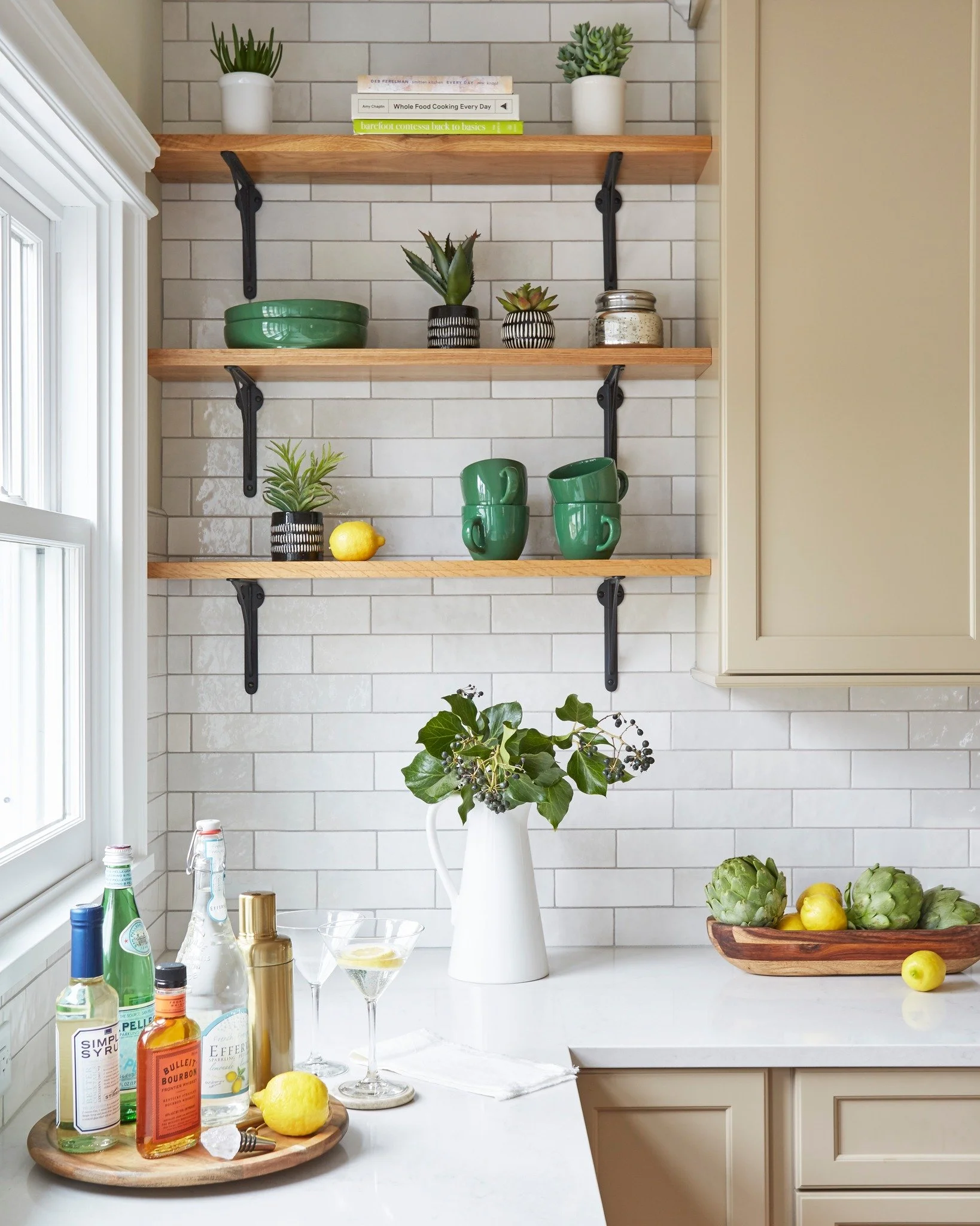 Kitchen with white subway tile backsplash and open wooden shelves holding potted plants, bowls, and cups, with a white countertop featuring a vase of greenery, assorted bottles, lemons, artichokes, and a martini glass.