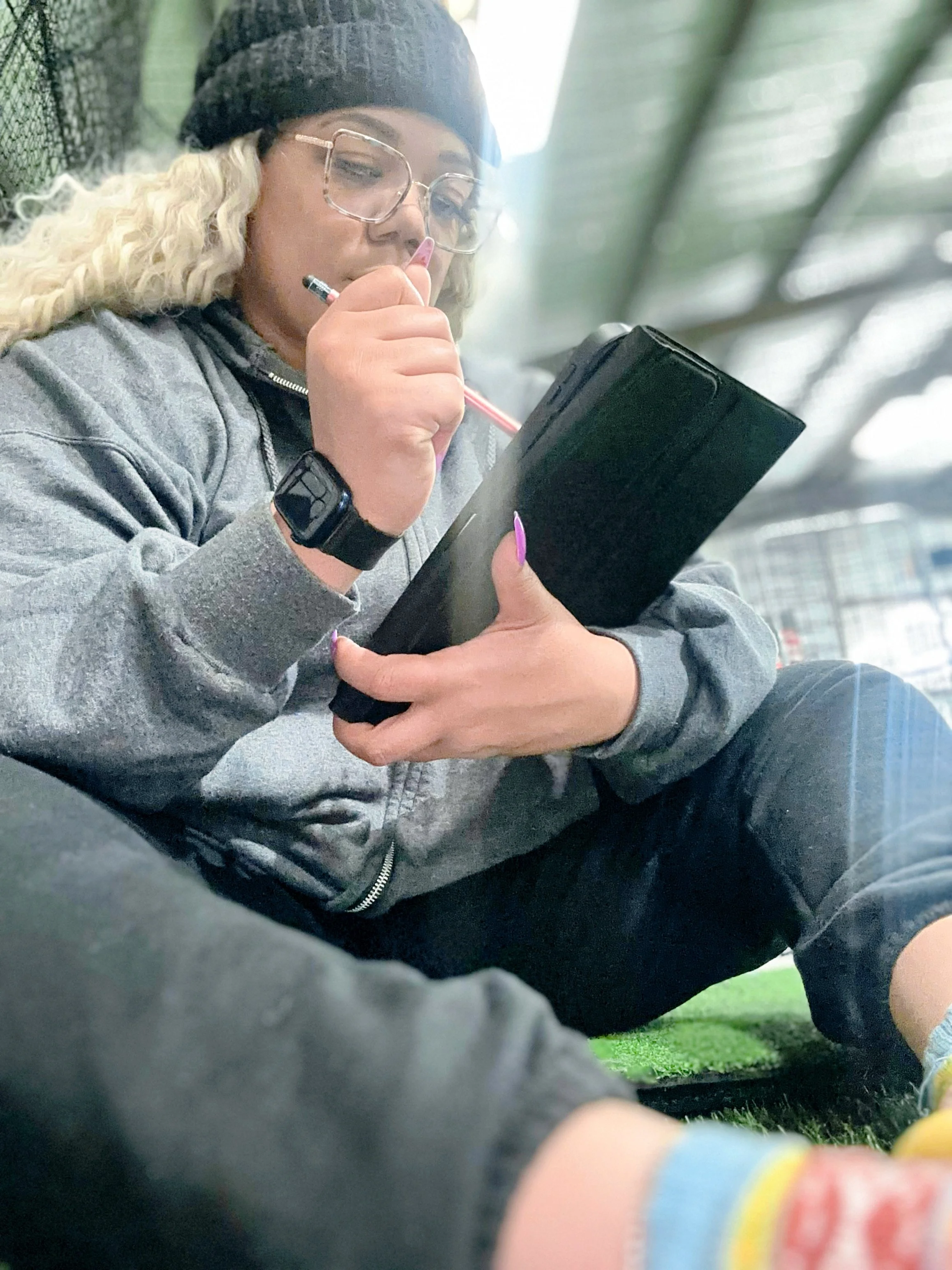 A young woman with long, curly blonde hair, glasses, a beanie hat, and a smartwatch, is sitting on the ground and writing in a black notebook with a pink pen. She is wearing a gray hoodie, black pants, and colorful socks, in a bright, outdoor setting with a green surface and a building in the background.