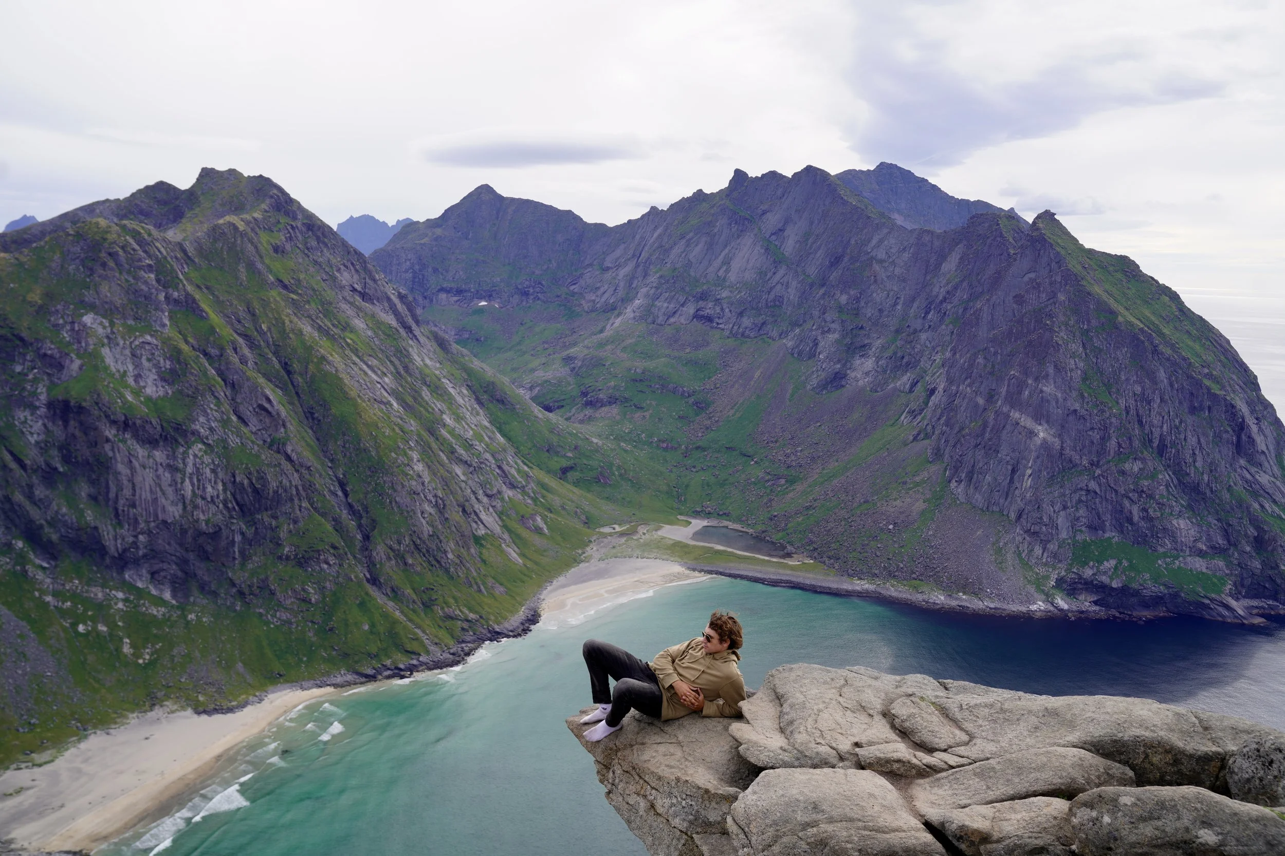 Person lying on large rock ledge overlooking a mountainous coastal landscape with green hills and a sandy beach below.