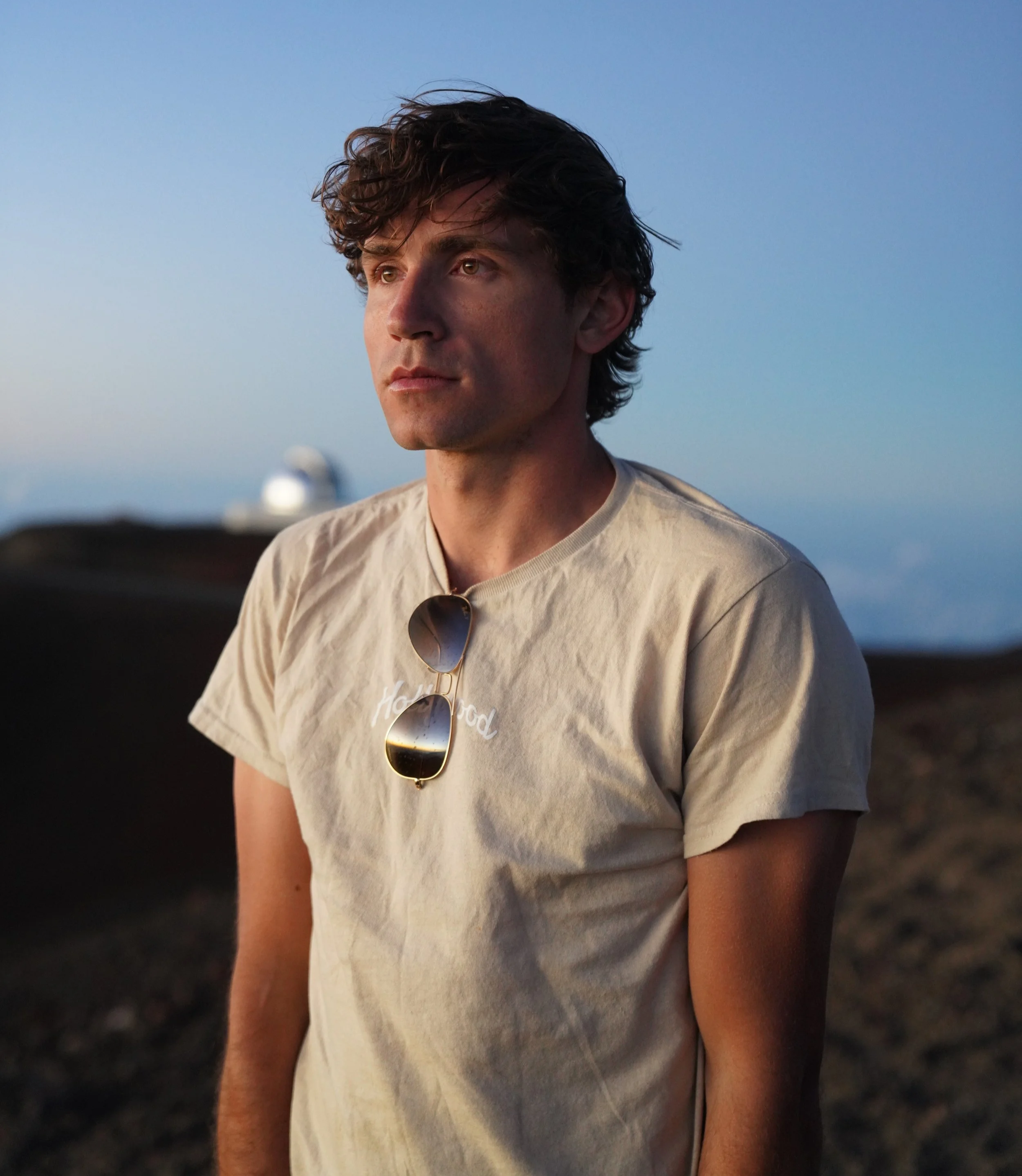 A young man with dark curly hair stands outdoors with a calm expression, wearing a beige T-shirt with a small white logo, and sunglasses hanging from the collar, during dusk or dawn.