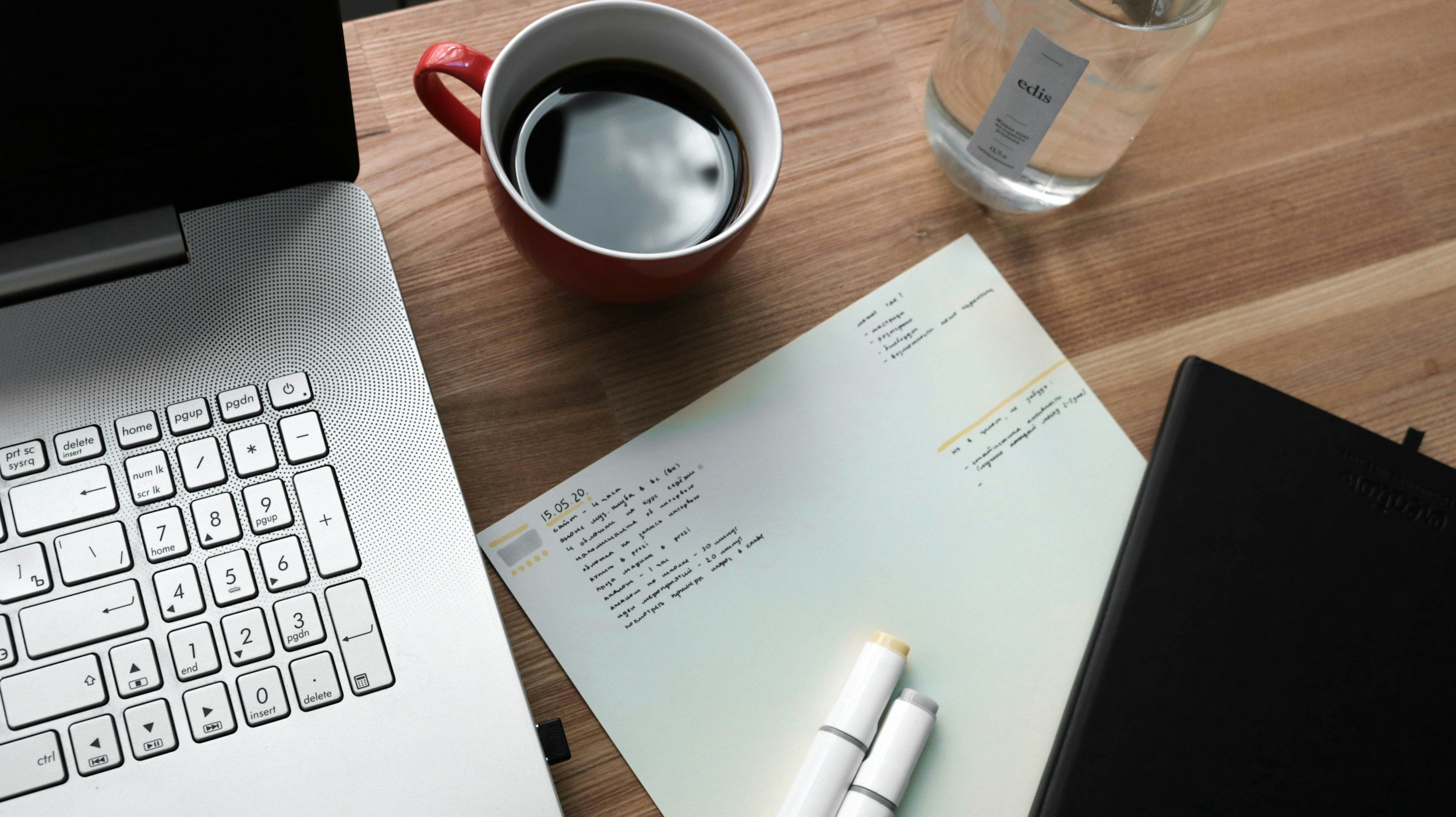 A workspace with a silver laptop, an open notebook with handwritten notes, a black notebook, a red coffee mug filled with black coffee, a bottle of water, and two white markers on a wooden desk.
