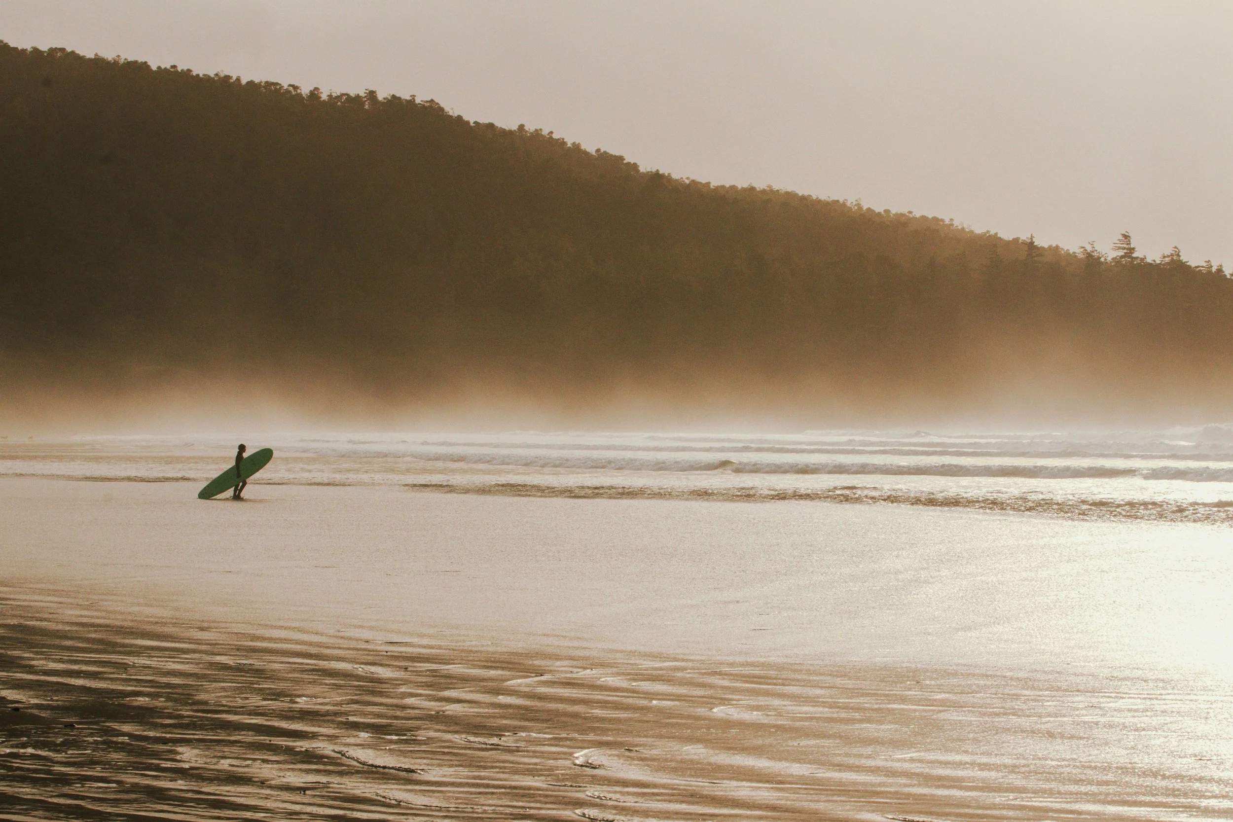A person holding a surfboard walking along the sandy beach at sunrise or sunset, with hills in the background and mist over the shoreline.
