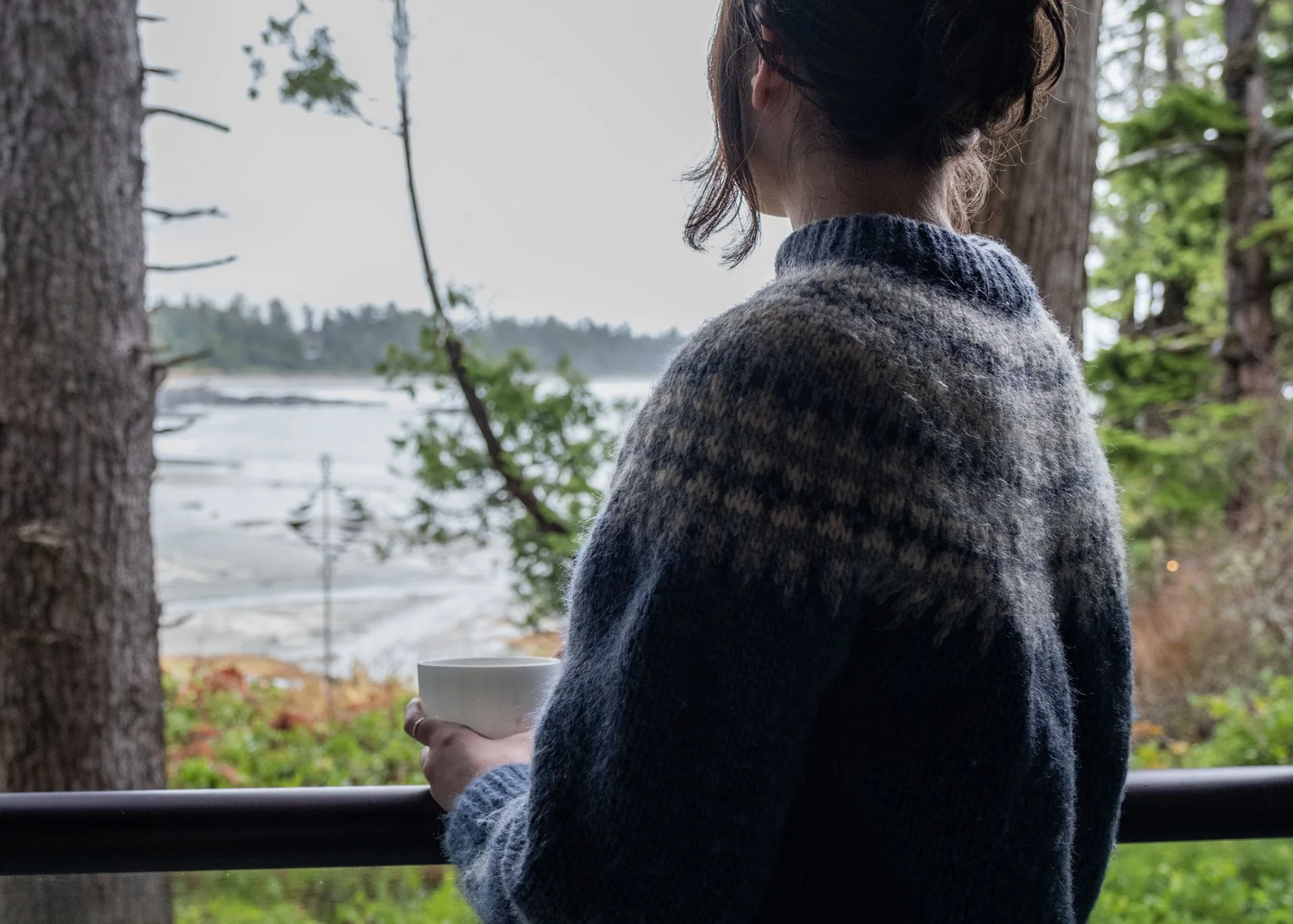 A woman with her back to the camera holding a white mug, looking out at a landscape of trees and a body of water on a cloudy day.