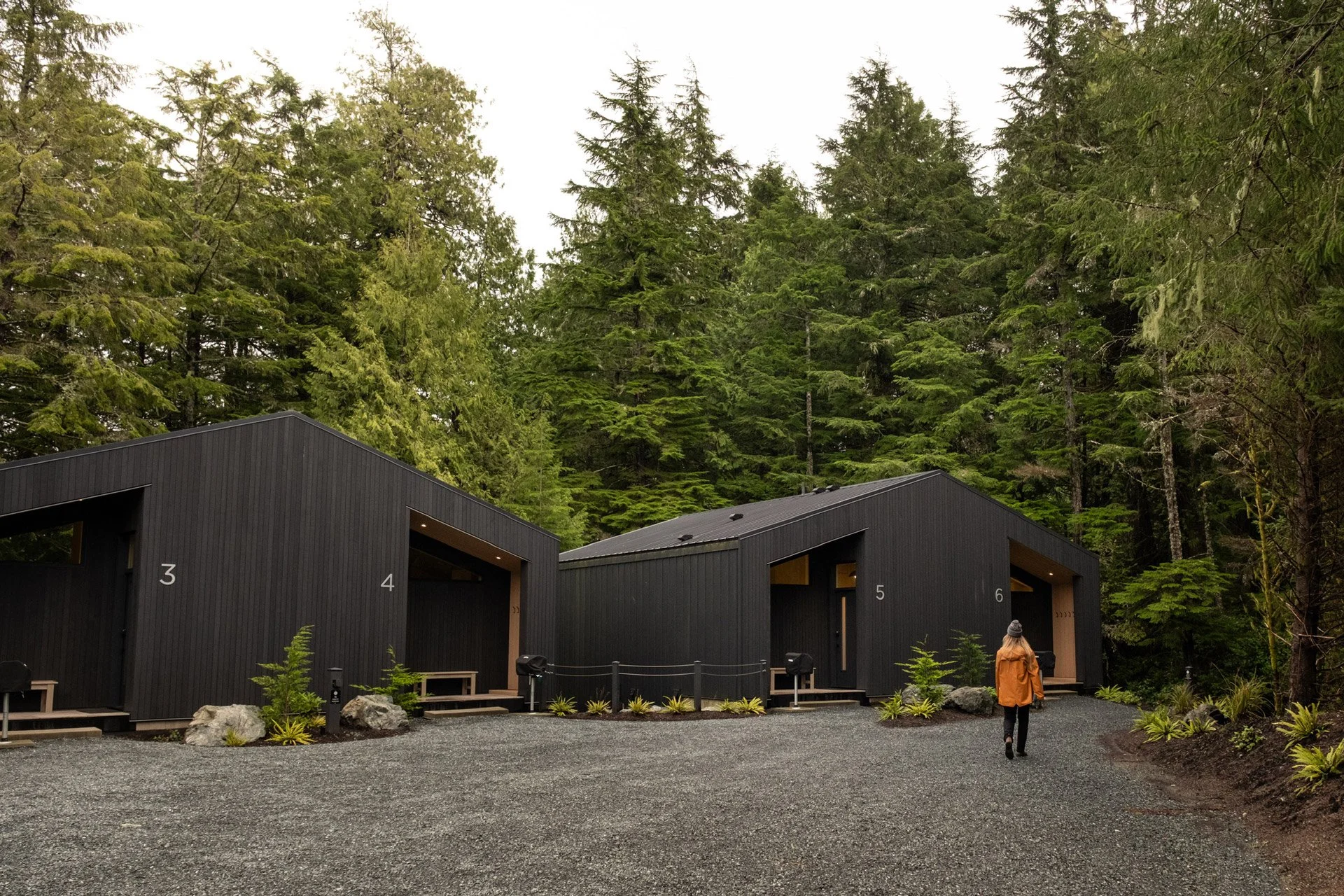 A person walks toward modern black cabins numbered 3, 4, 5, and 6, set in a forested area with tall evergreen trees and cloudy sky.