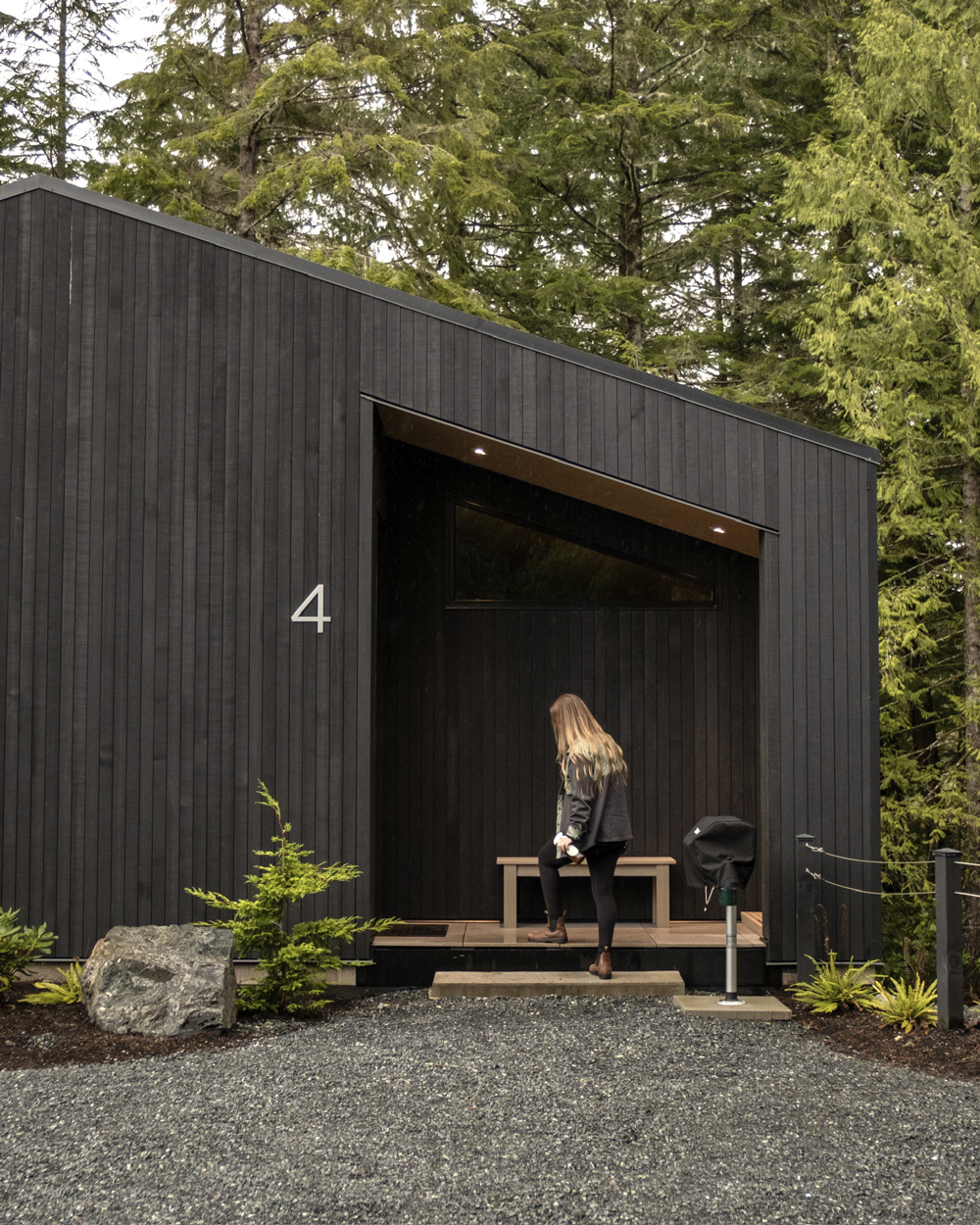 A modern black house with the number 4 on the exterior, featuring a small porch with a bench, surrounded by trees and greenery.