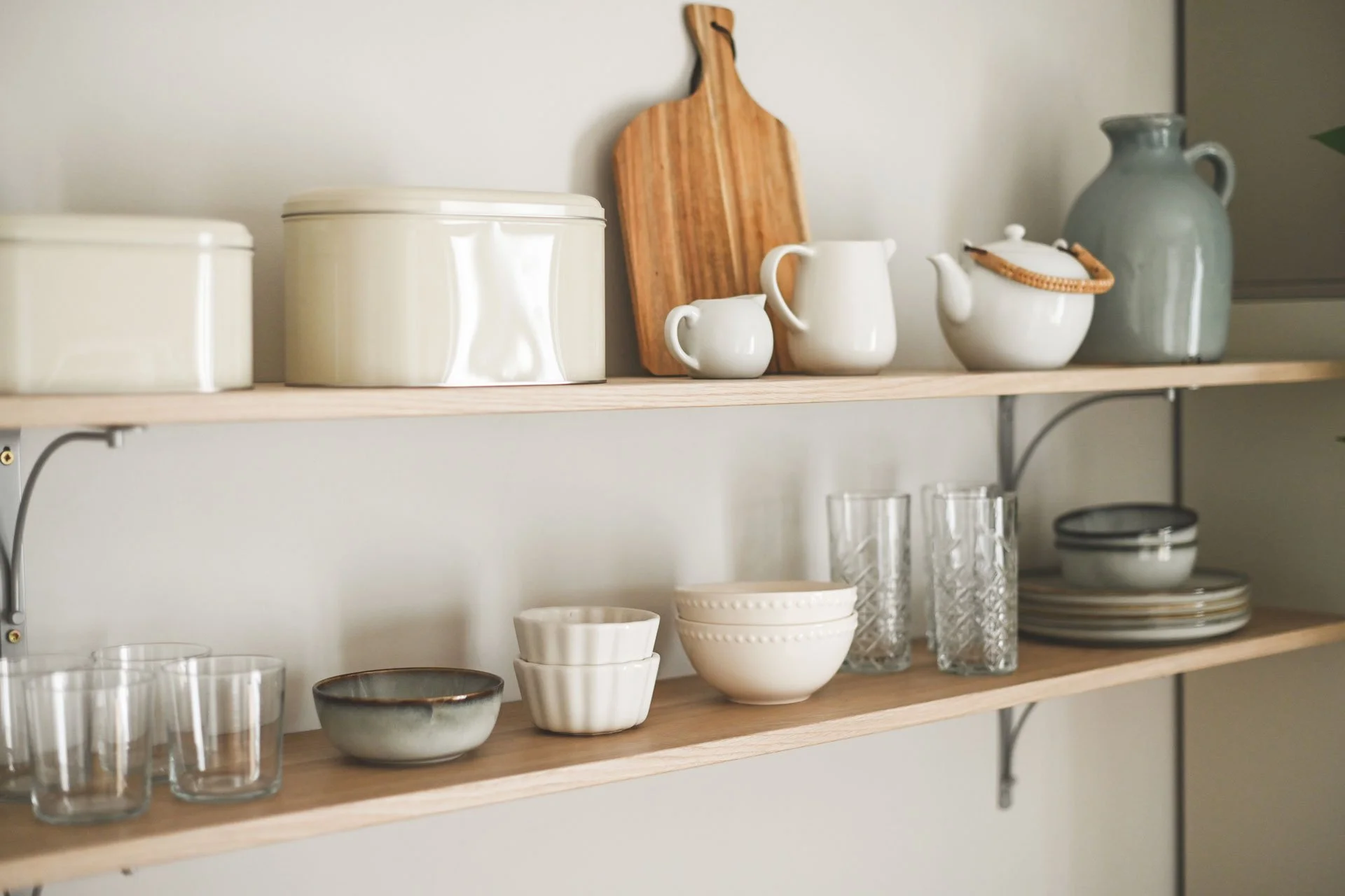 Kitchen shelves holding ceramic and glassware, including bowls, cups, a pitcher, a teapot, and a wooden cutting board.