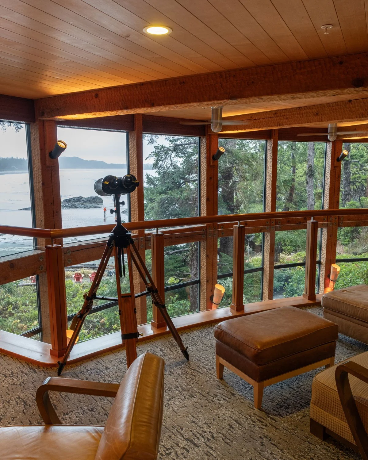 Indoor wooden room with large windows overlooking a beach and forest, telescope on tripod, leather armchair, ottoman, and cozy lighting.