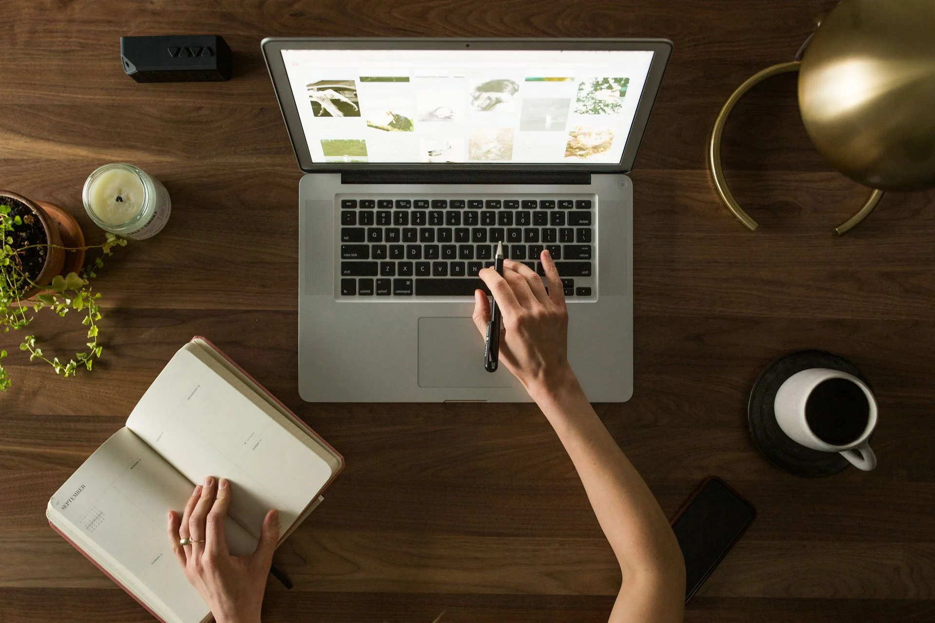 Top-down view of a wooden desk with a person using a laptop, a notebook, a cup of coffee, a candle, a plant, a small speaker, a smartphone, and a gold table lamp.