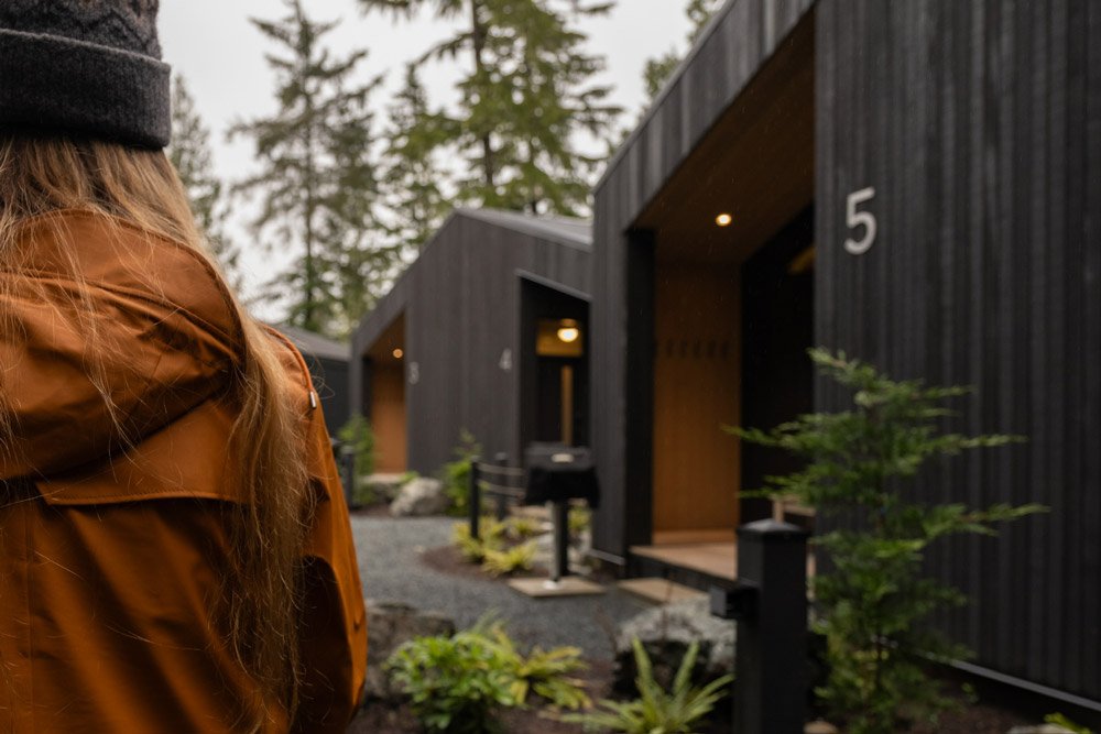 A person with long hair wearing a brown jacket and dark beanie looking at several dark-colored cabin units with large numbers 4 and 5, surrounded by trees and outdoor landscaping.