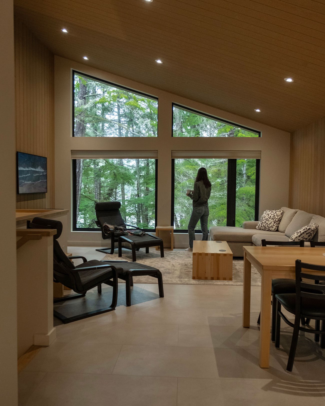 Living room with large windows showing trees outside, a woman standing and looking out, and various chairs and a sofa.