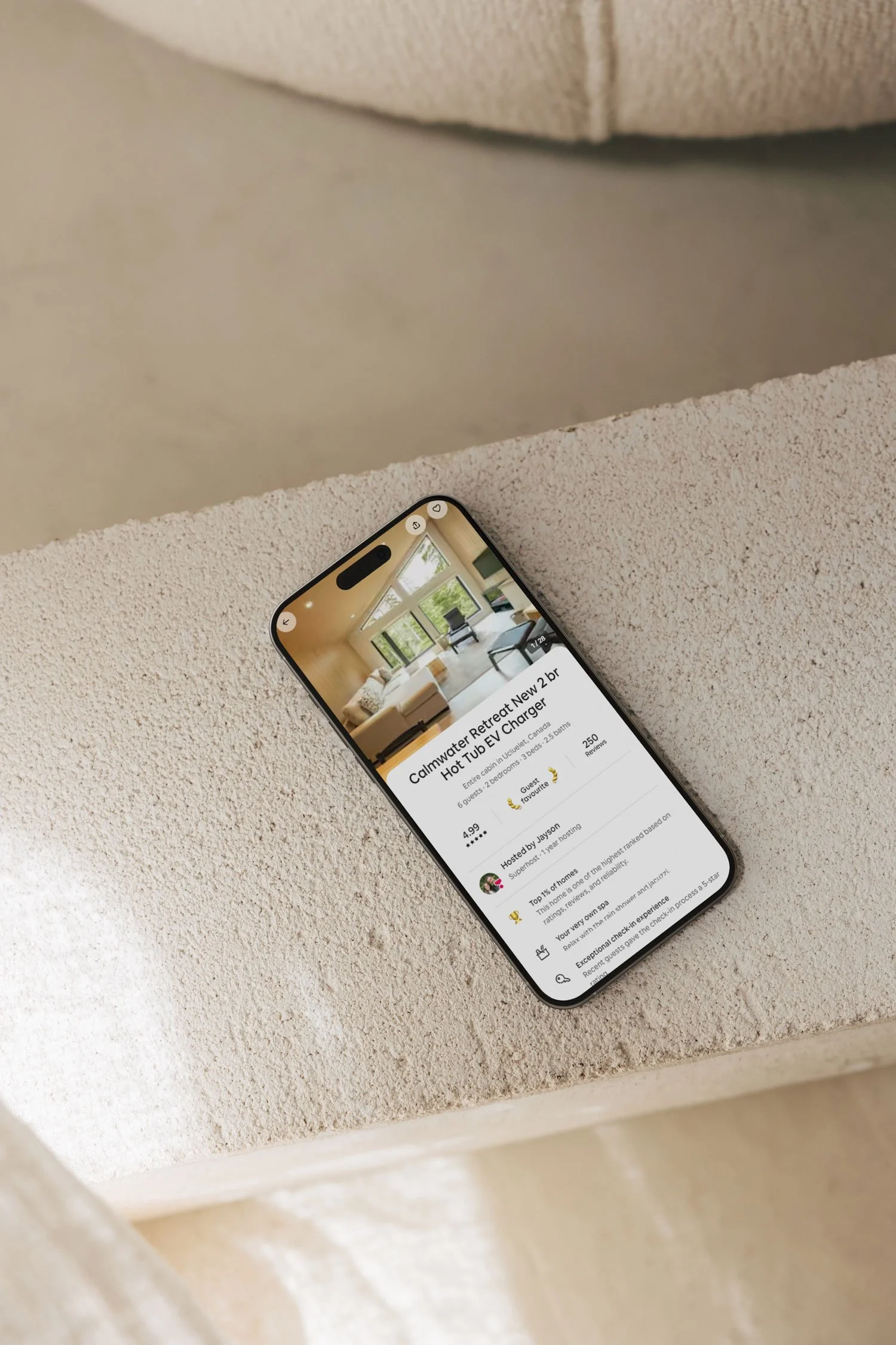 Smartphone on a concrete ledge displaying a booking for Calmr Retreat New 2br Hot Tub EV Charger, with an interior room image and booking details visible.