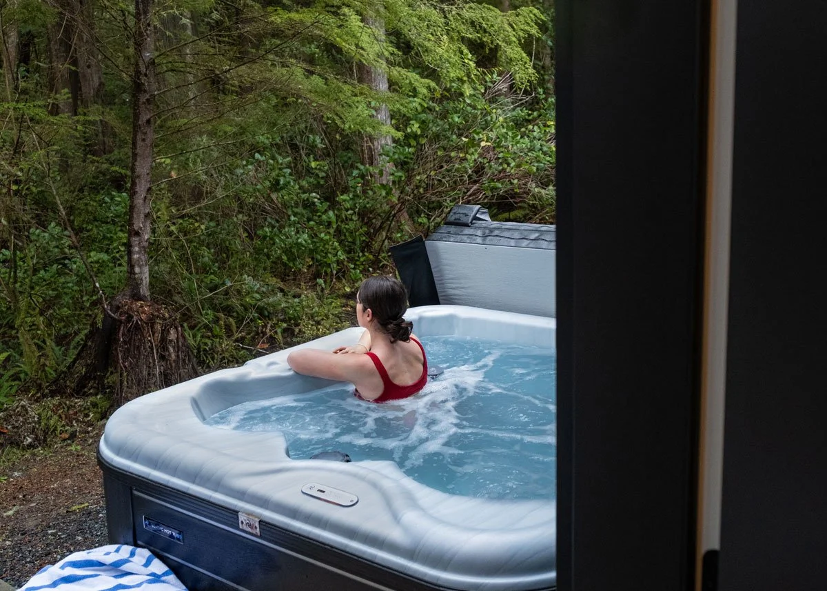 A woman in a red swimsuit relaxing in an outdoor hot tub surrounded by trees and greenery.