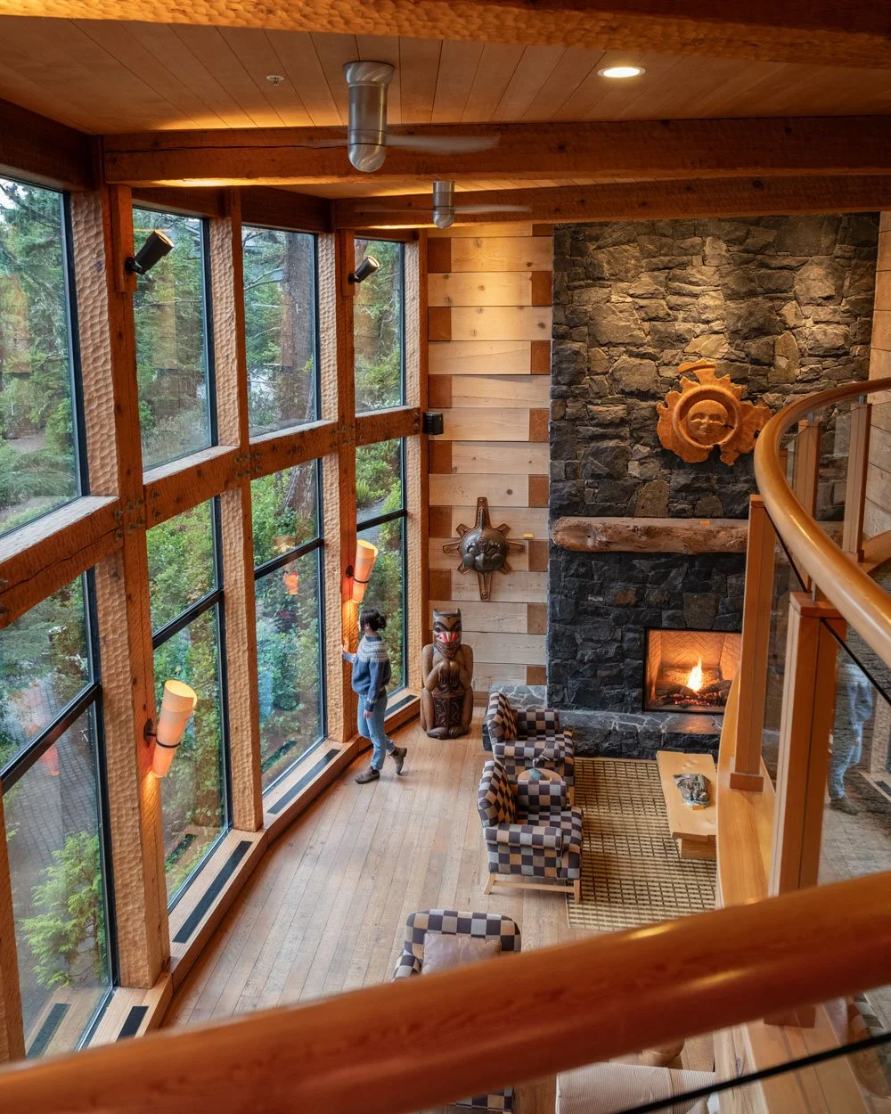 A cozy living room with a high wooden ceiling, large curved windows, a stone fireplace with a fire, and Native American art and masks on the walls. A person stands near the window, looking outside.