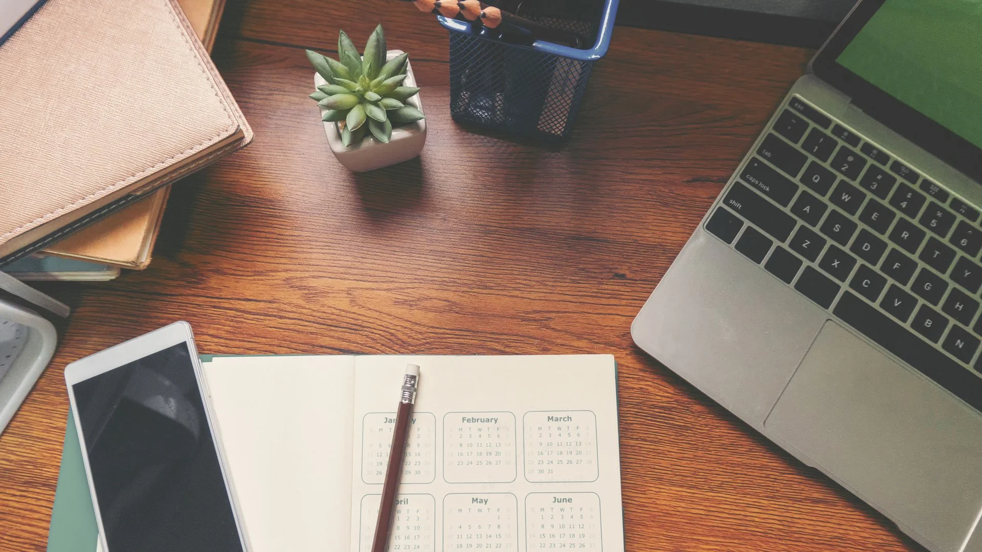 A wooden desk with a laptop, a smartphone, a calendar, a plant, and office supplies.