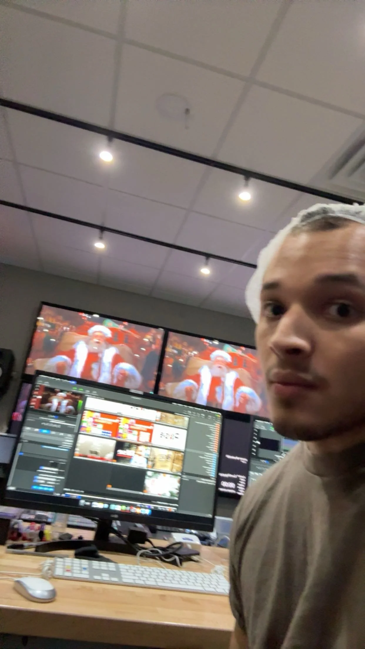 A man in a brown shirt and wearing a white hairnet takes a selfie in a control room with dual monitors displaying video editing software. In the background, two screens show Santa Claus.