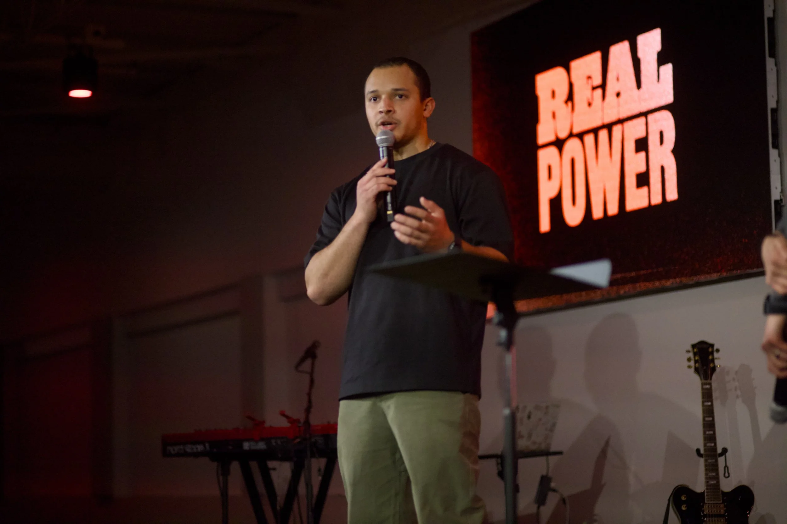 A man in a black shirt and tan pants speaks into a microphone on stage, with a large screen behind him displaying the words 'REAL POWER' in red letters.