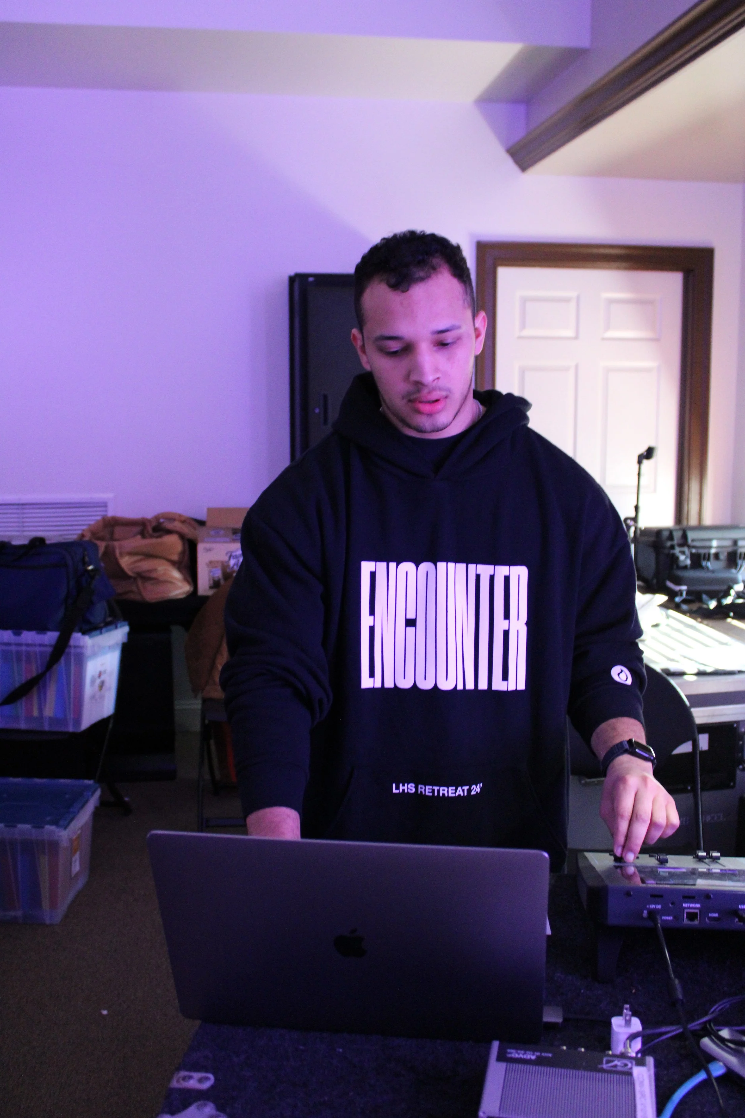 A young man with short dark hair and light skin working at a DJ setup, wearing a black hoodie with the words "ENCOUNTER" and "LHS RETREAT 24" printed on it, in a room with purple lighting.