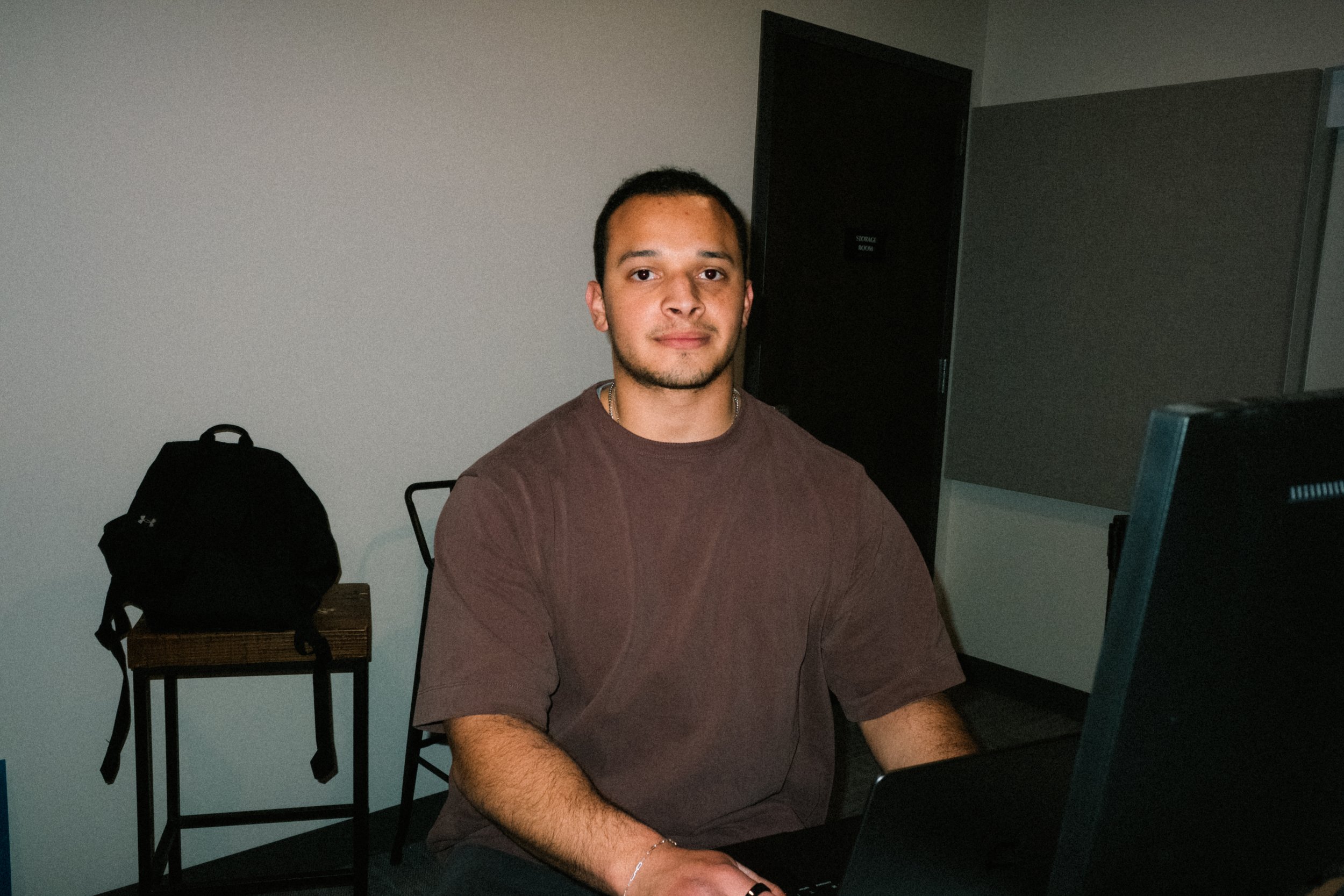 A young man sitting at a table with a laptop in front of him, in a room with plain walls and a closed door, with a backpack on a chair behind him.
