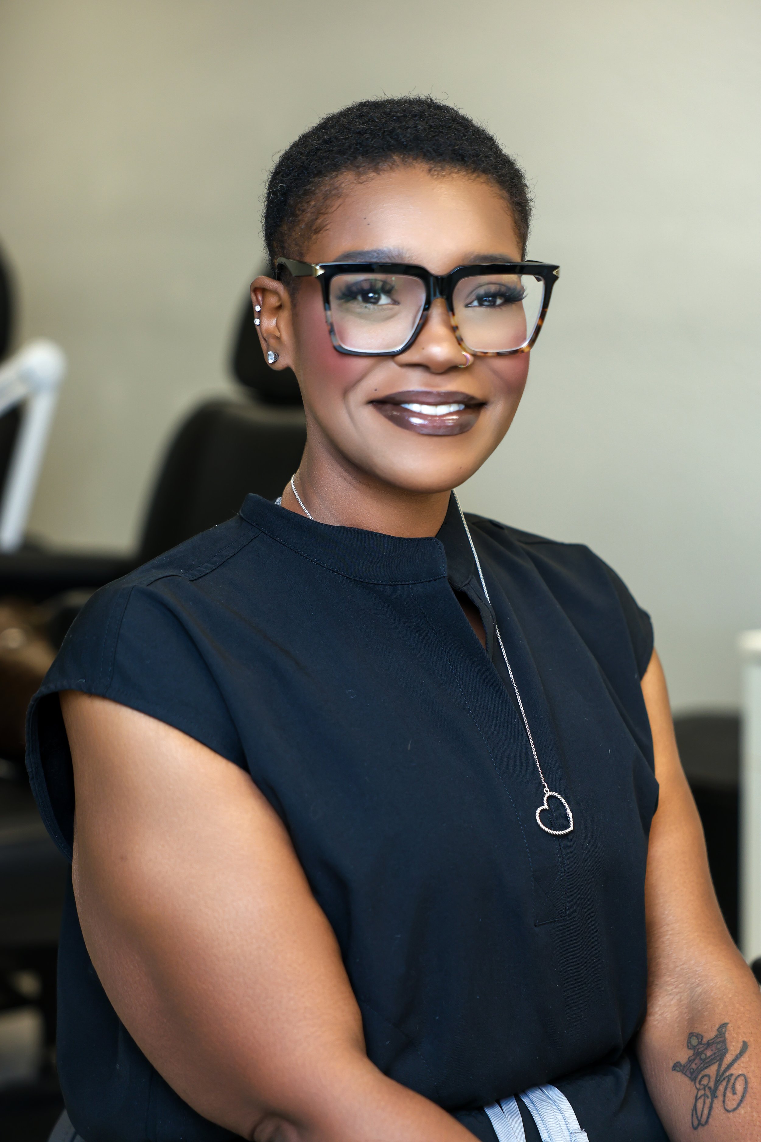 A smiling woman with short hair, wearing large glasses, a dark top, and a heart-shaped necklace, sitting in an office setting.
