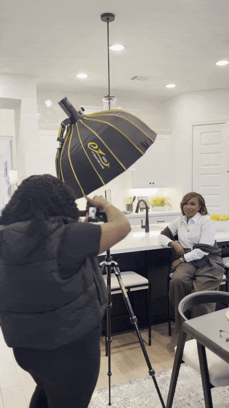A woman sitting in a kitchen position, posing for a photoshoot with a professional photographer shooting her. The photoshoot is happening indoors with bright ceiling lights, and the background shows a modern kitchen with minimalist decor.