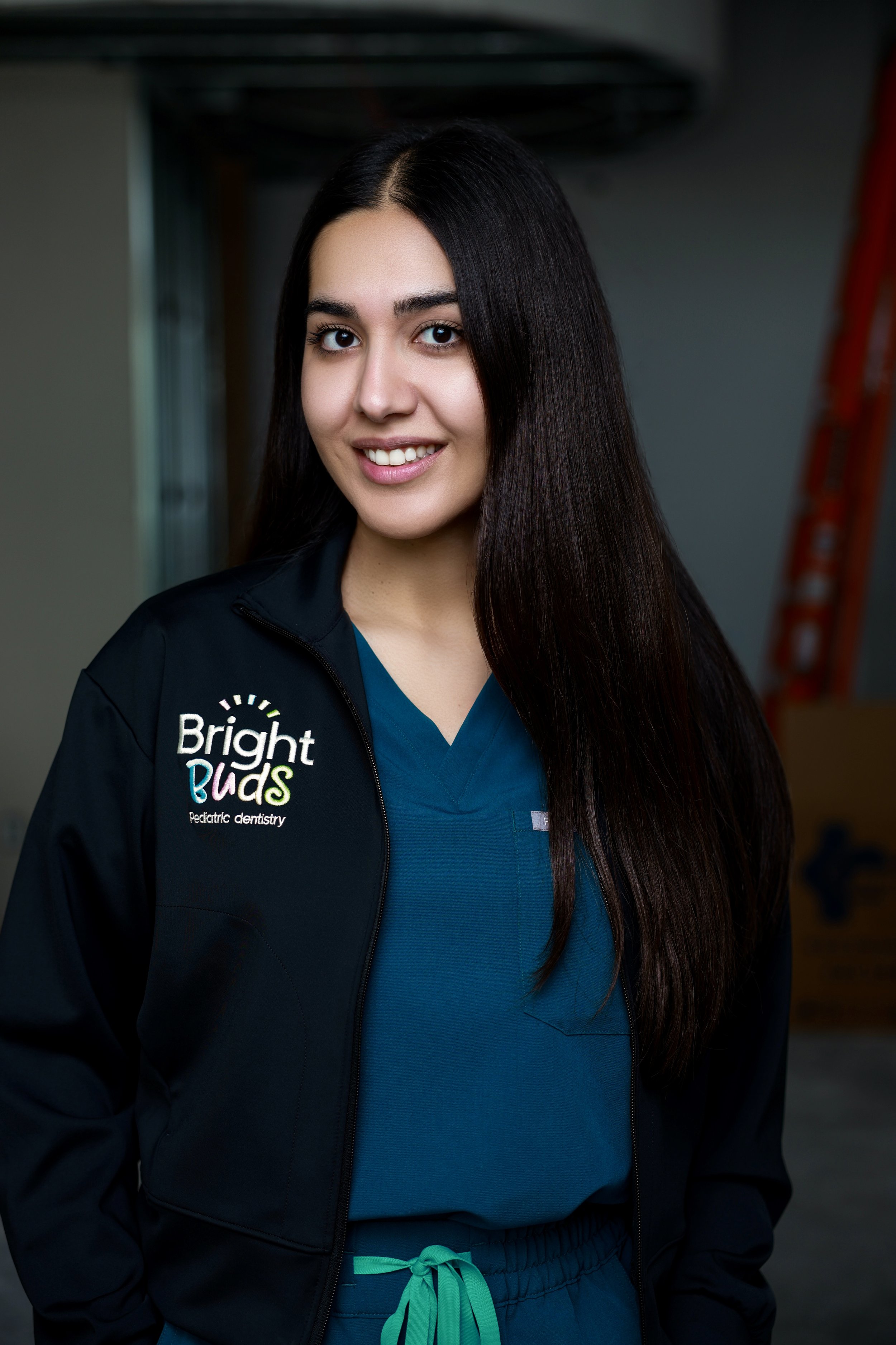 A young woman with long dark hair wearing blue medical scrubs and a black jacket with 'Bright Buds Pediatric Dentistry' embroidered on it, smiling at the camera.