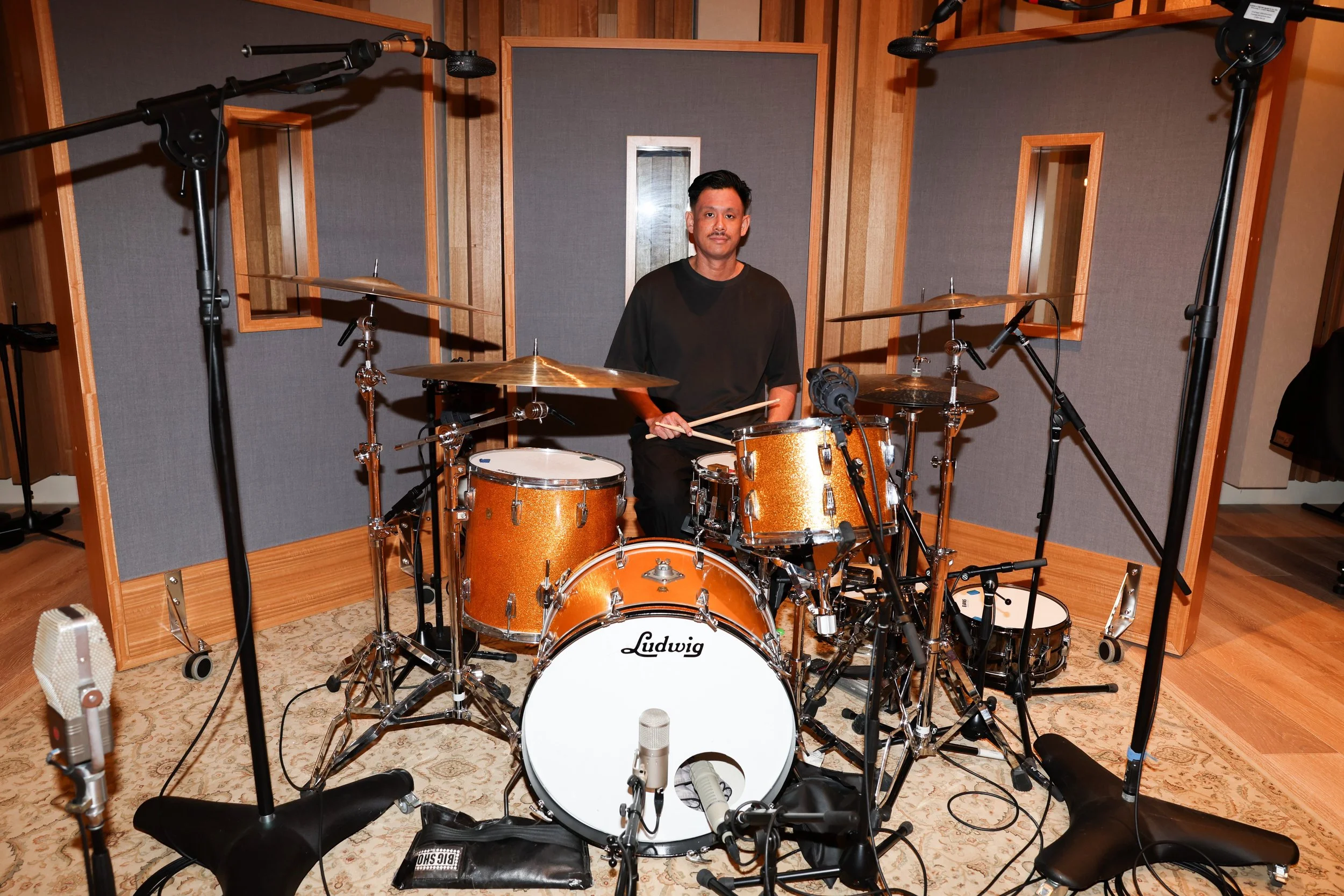 A man sitting behind a Ludwig drum set in a recording studio with soundproof walls and multiple microphones surrounding him.