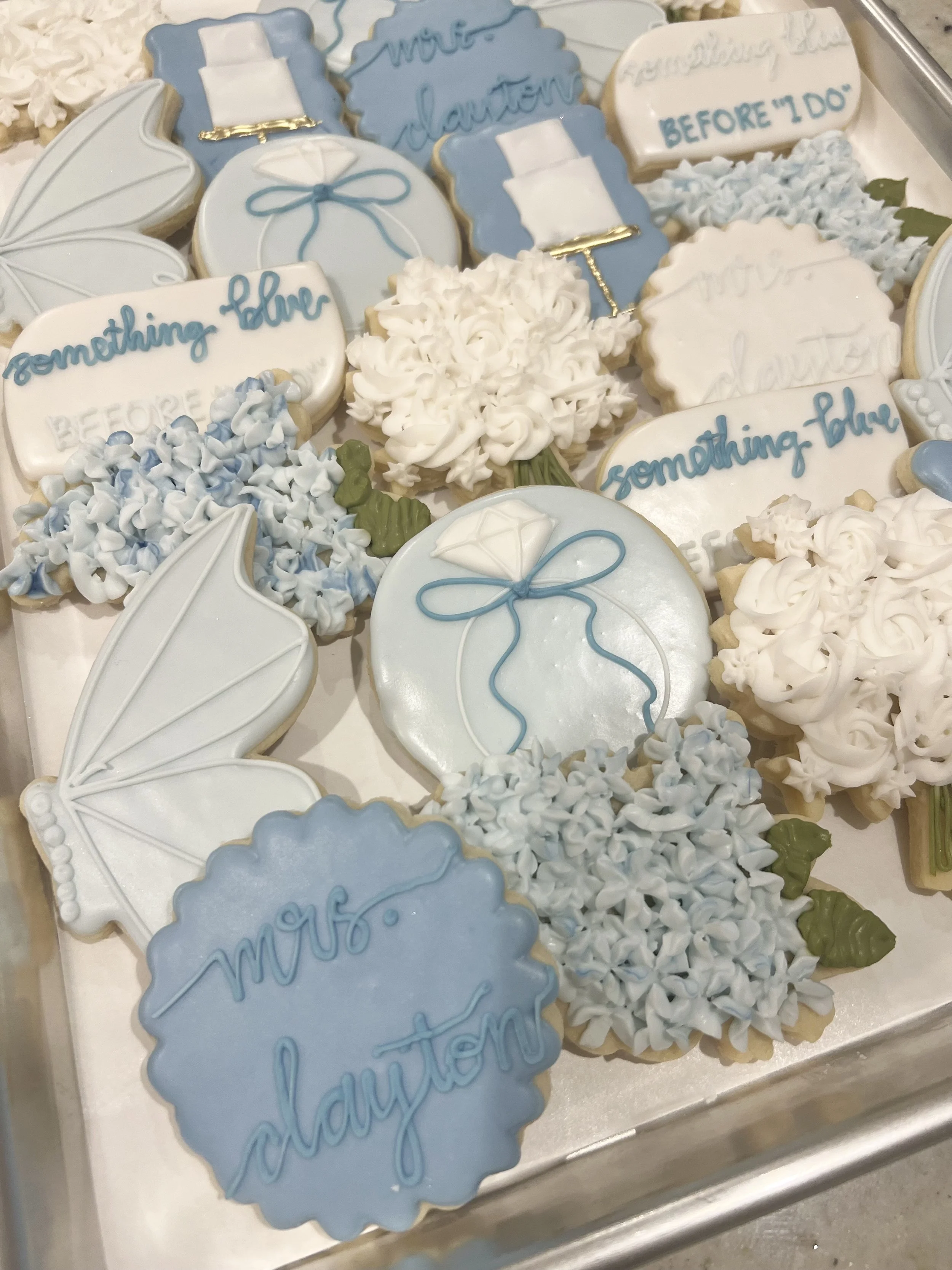 Assorted decorated cookies with blue, white, and green icing, featuring phrases like "Mrs. Clayton" and "something blue," along with floral, umbrella, and gift box designs.