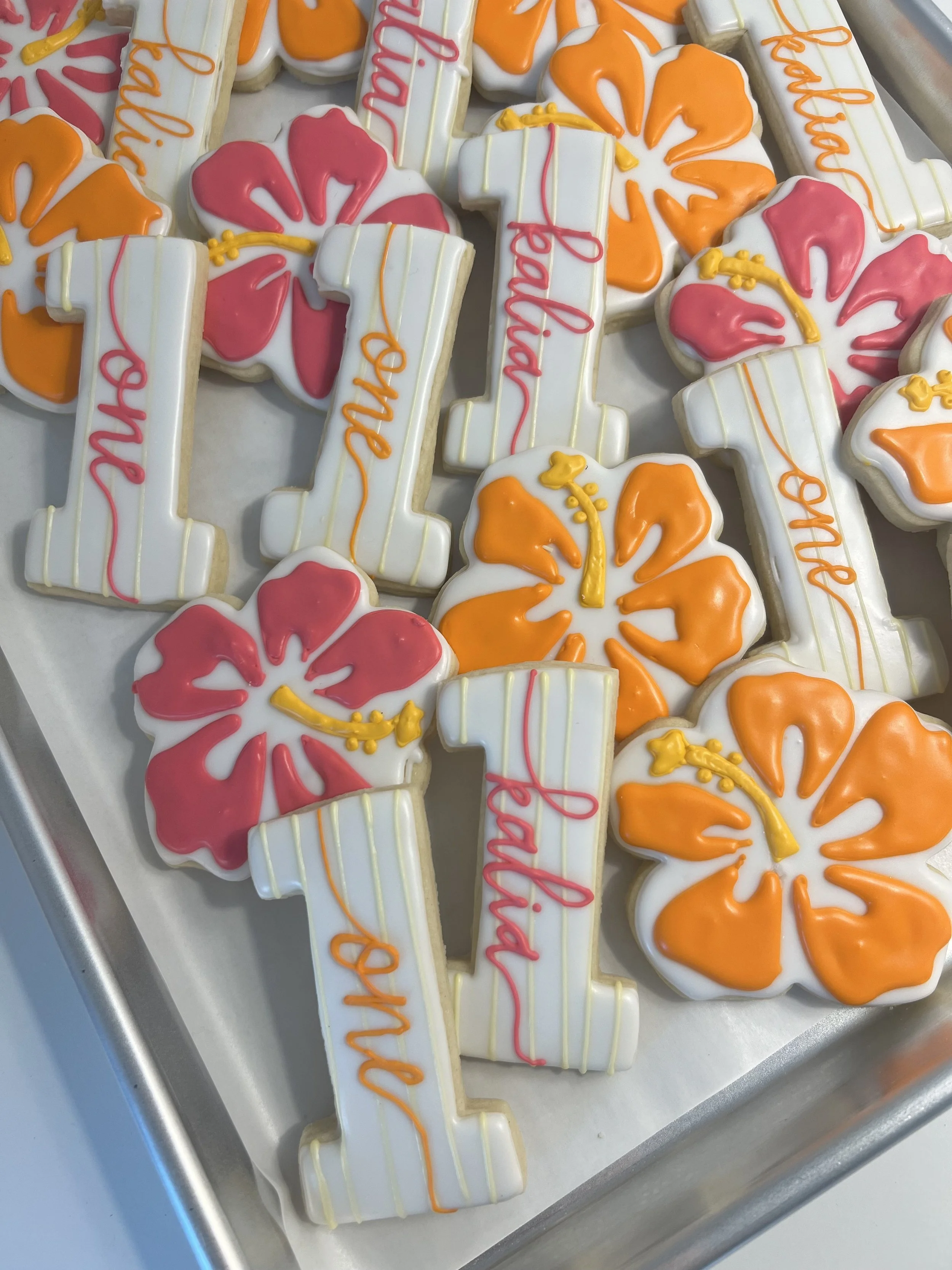 Decorated cookies shaped like the number one and hibiscus flowers, with pink, orange, and yellow icing, placed on a white tray.