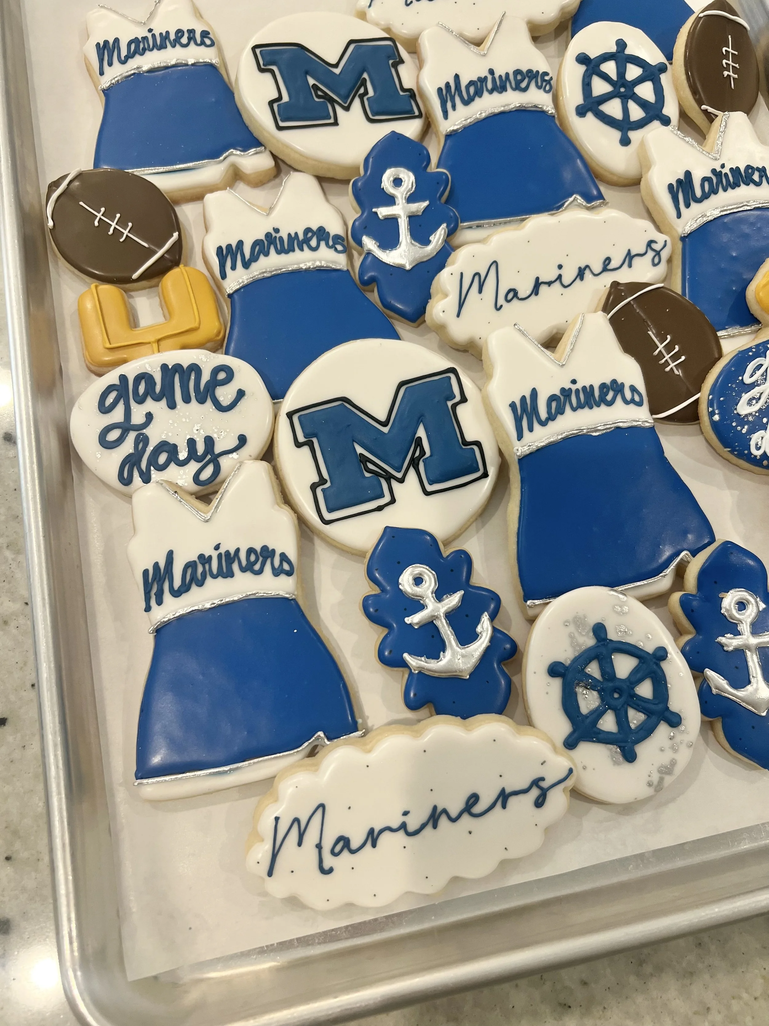 Tray of decorated cookies with a nautical and sports theme in blue, white, and brown. Some cookies feature the word 'Mariners' and a large blue 'M' logo. Others depict anchors, footballs, the number 4, and ship wheels.