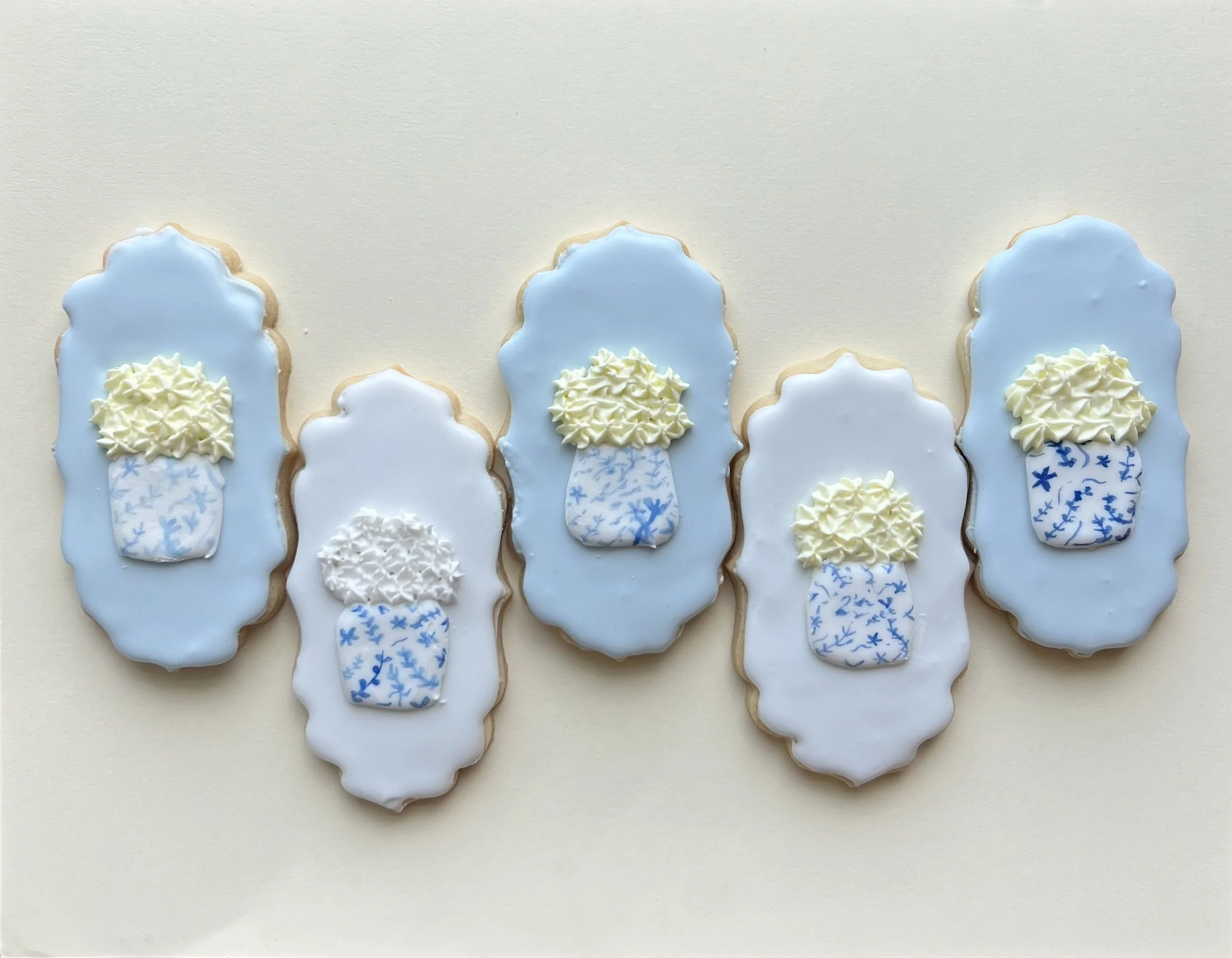 Five decorated cookies with blue and white theme, each shaped like a vertical oval with scalloped edges, topped with icing and a small snowflake-patterned snowball decoration in a blue and white container.
