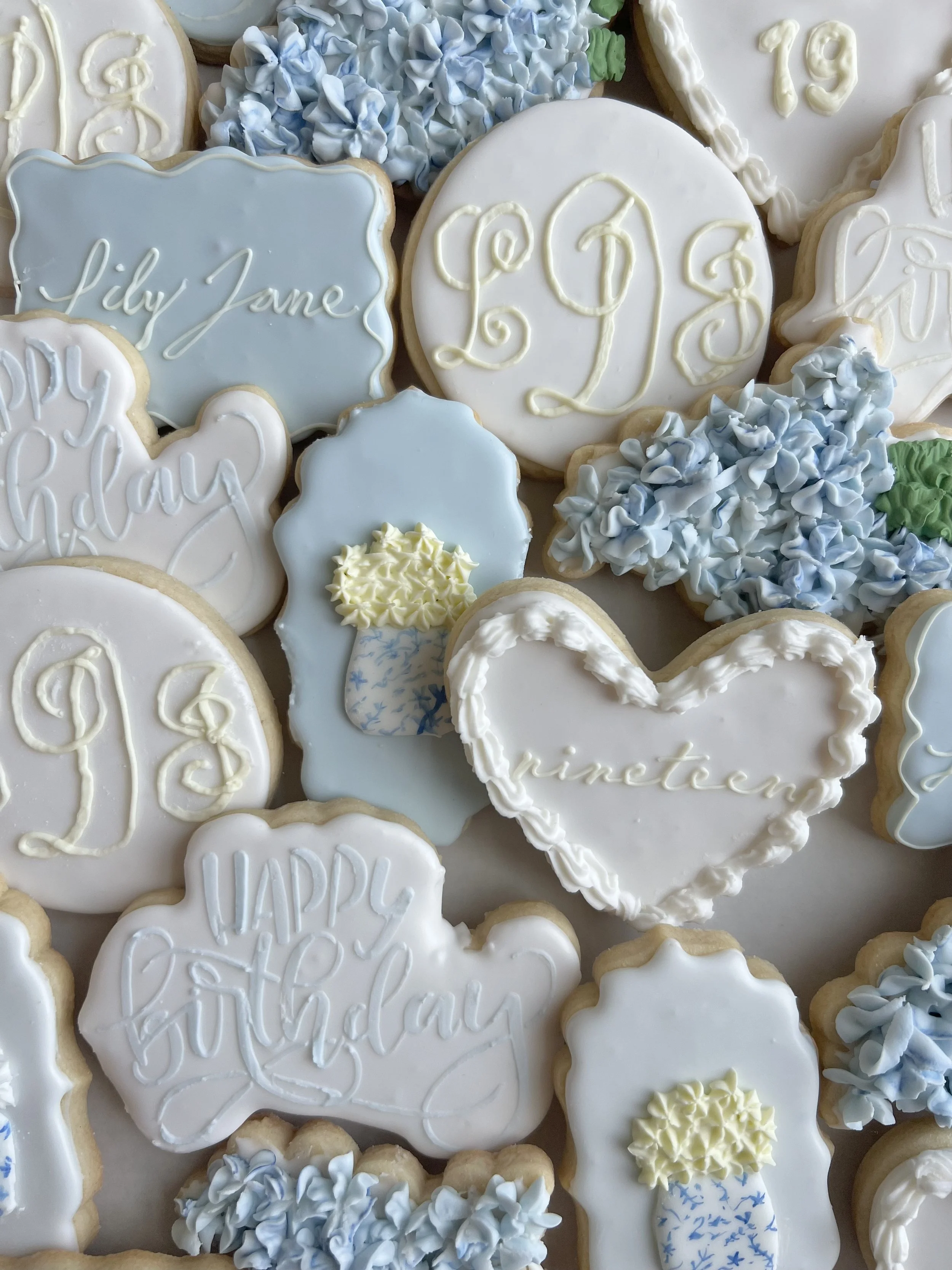 Decorated sugar cookies with baby shower and birthday themes in white, blue, and yellow icing featuring letters, flowers, a heart, and a bottle.