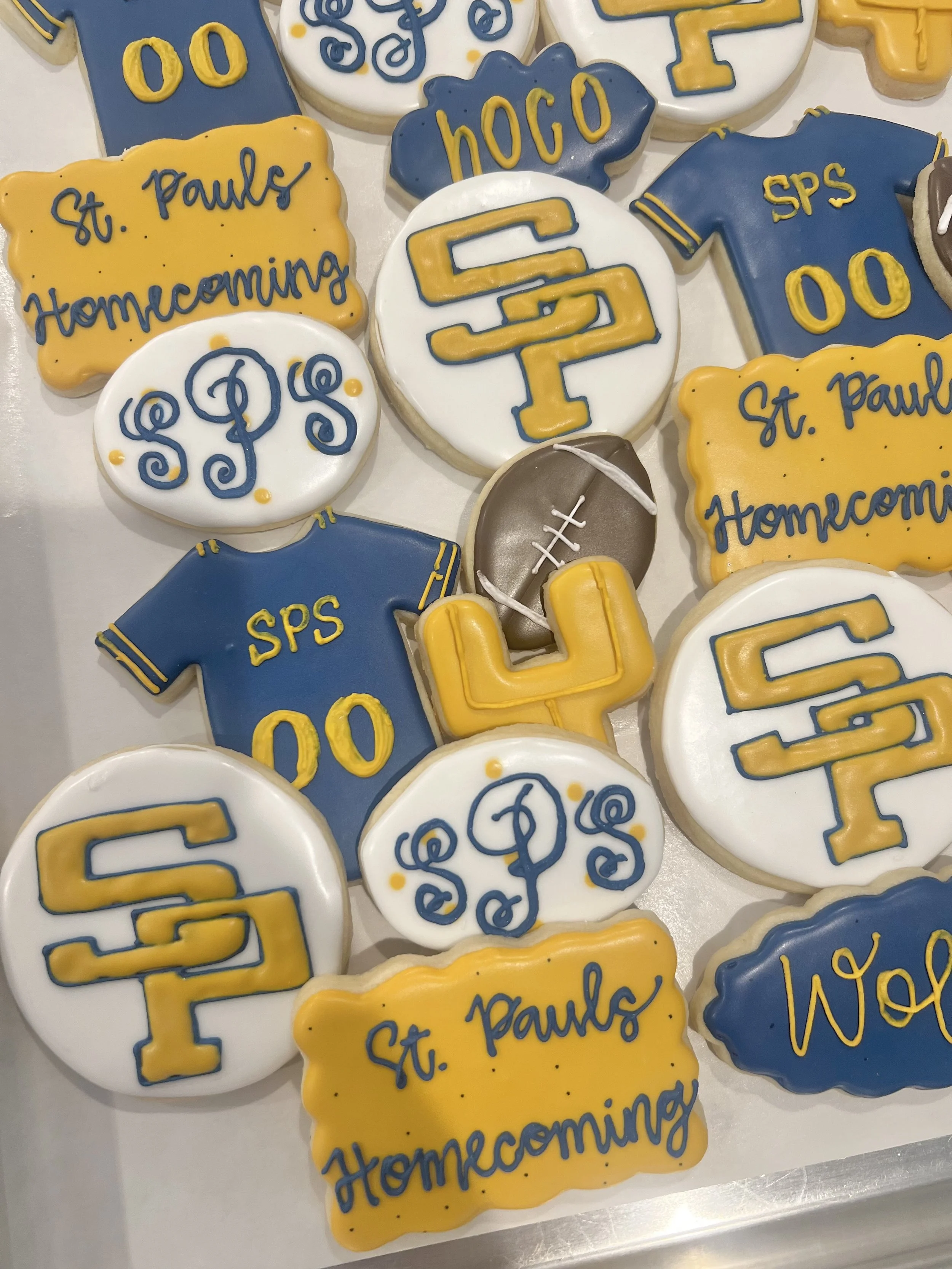 Assorted cookies decorated with blue, yellow, and white icing, featuring phrases like 'St. Pauls Homecoming', and symbols like footballs and the initials 'SPS' and 'PSC' for St. Pauls School, likely for a homecoming celebration.