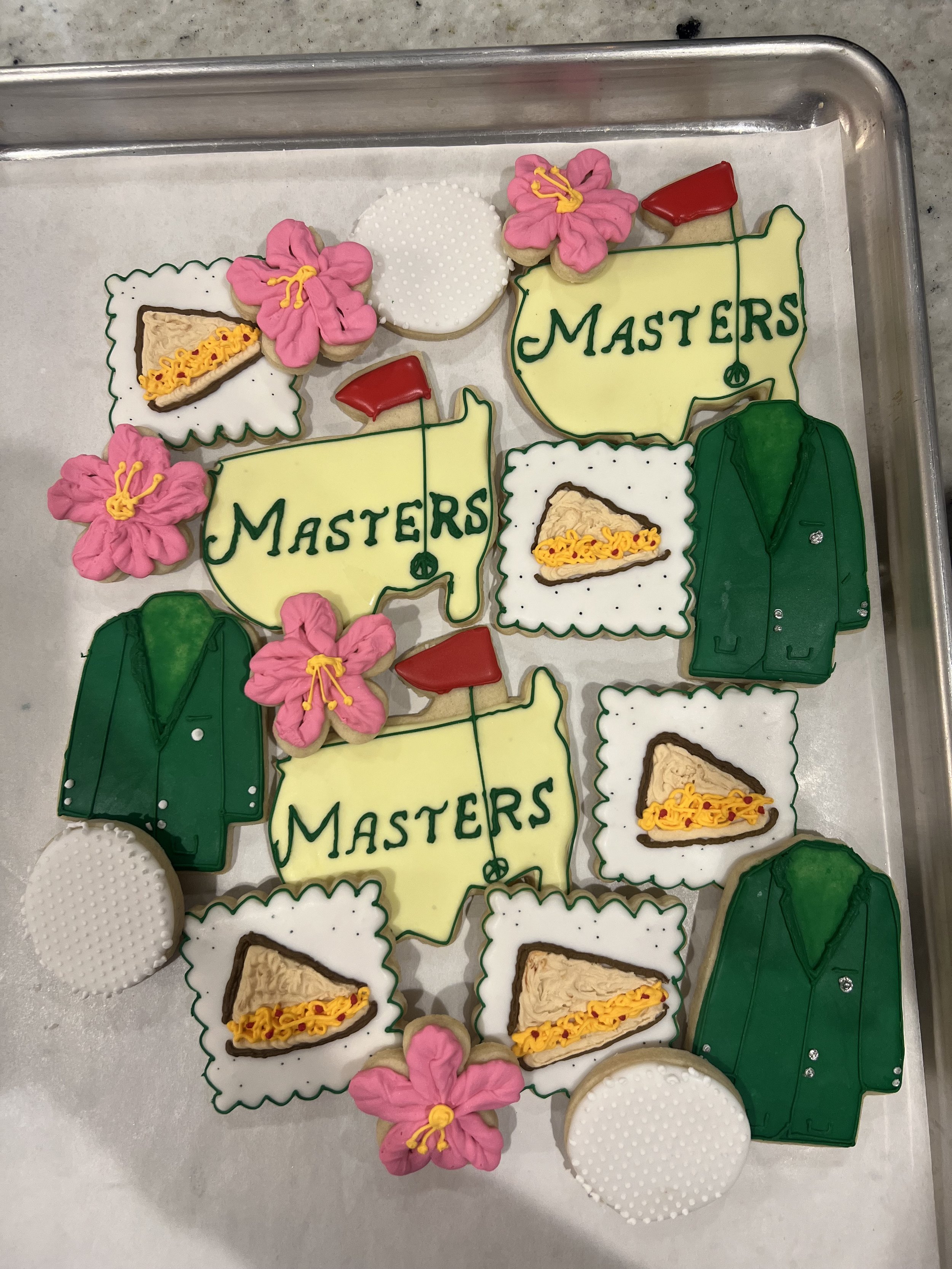 Assorted decorated cookies on a tray, including three cookies shaped like the state of Texas with 'MASTERS' written on them, three cookies shaped like green jackets, three cookies with pink flowers, three white cookies with textured patterns, and thr