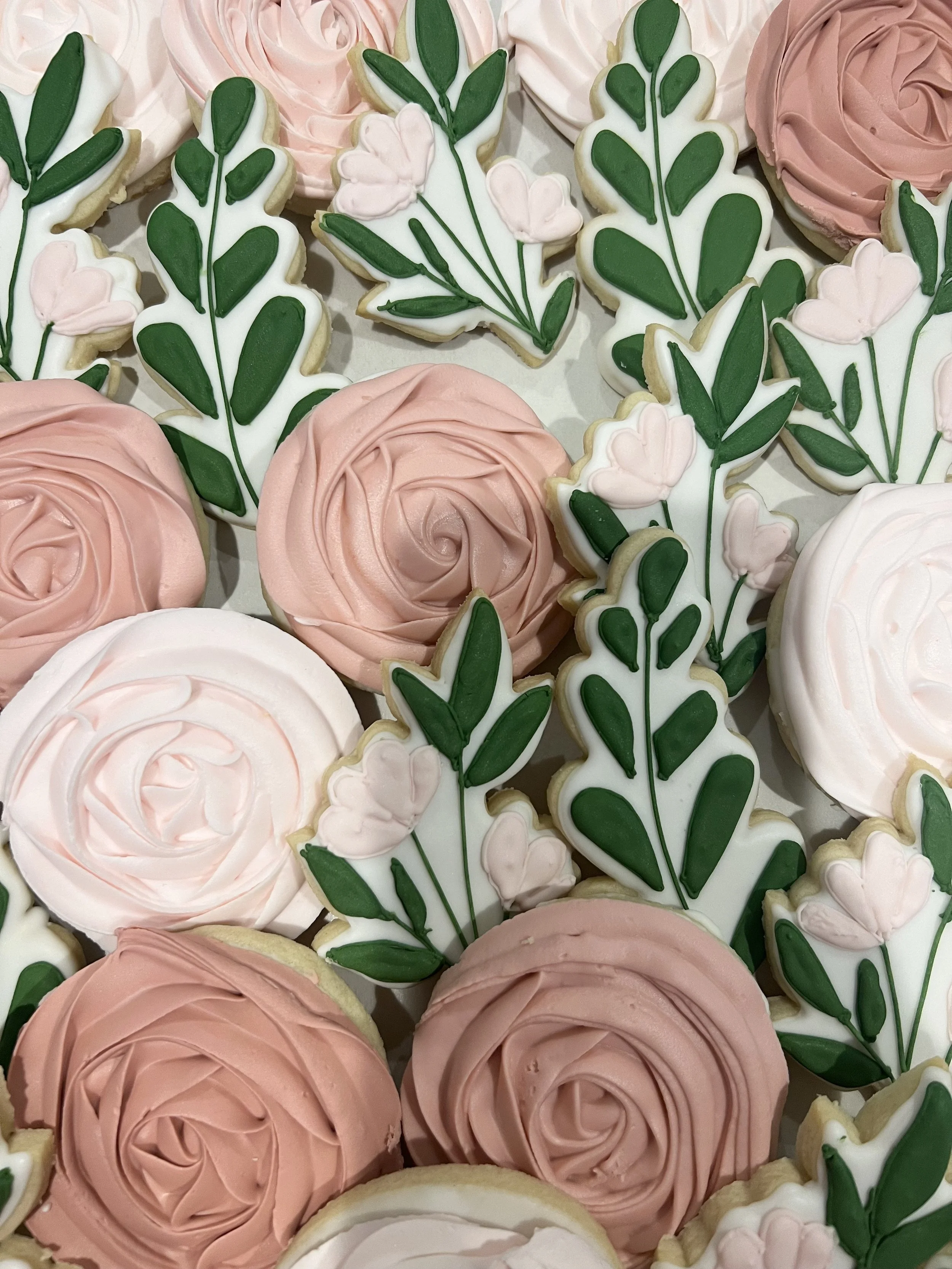 Decorative floral cookies in pink and white swirls, shaped like roses, and green leaf and pink flower-shaped cookies with detailed icing decorations.