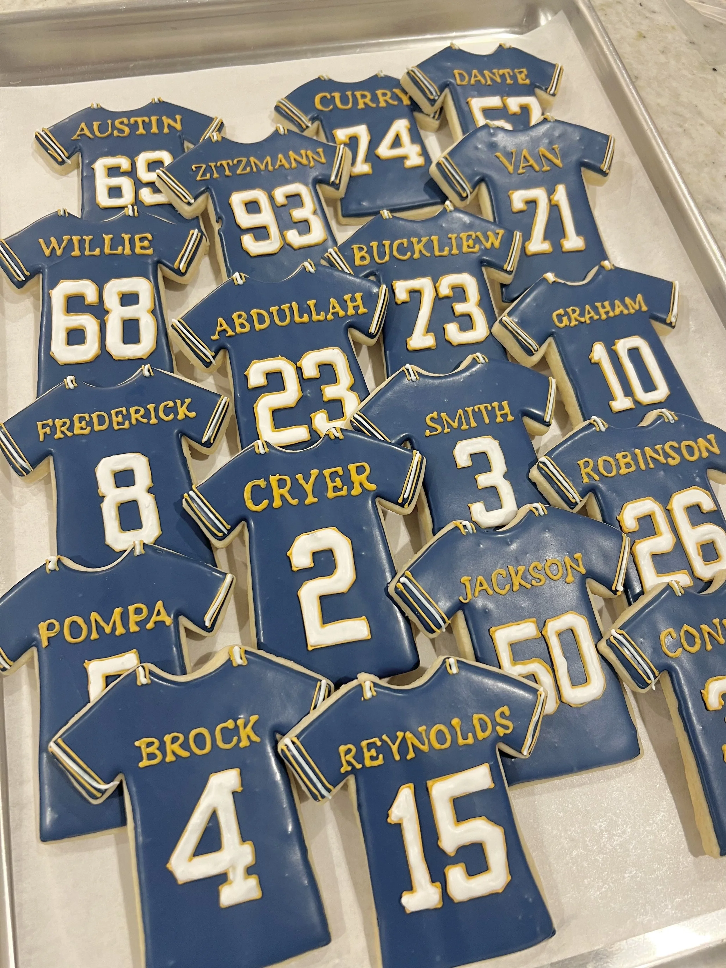 Decorated Mandeville High School Skippers football-shaped cookies with blue icing, yellow piping, and player names and numbers painted on them.