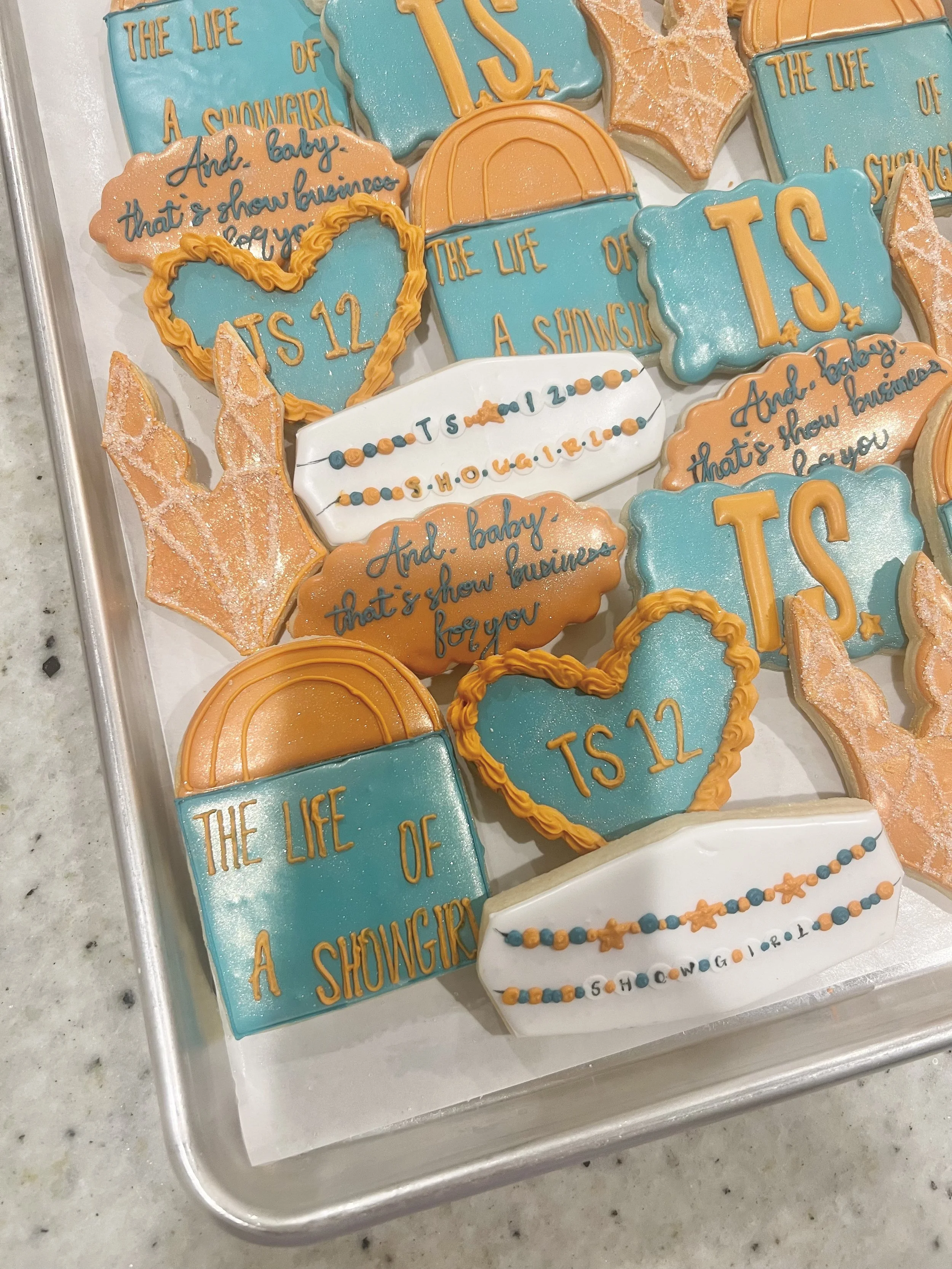 Decorated cookies celebrating a show titled 'The Life of a Showgirl,' with themes related to TS 12, birthdays, and showbiz. The cookies are in various shapes, including hearts, stars, and speech bubbles in orange, blue, and white colors.